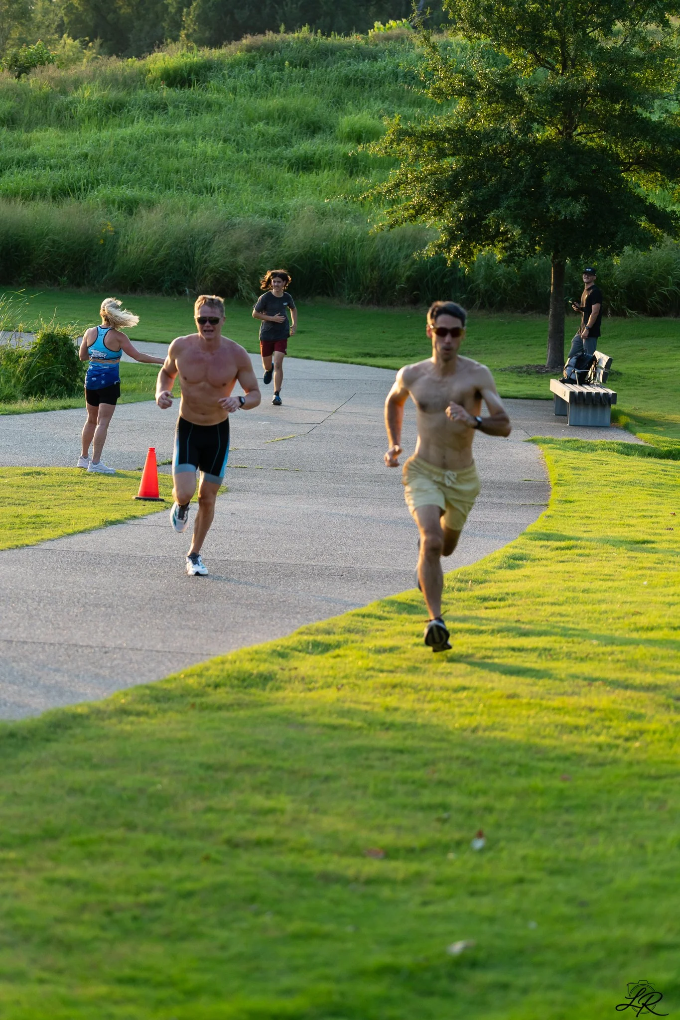 People running in a park on a paved pathway with grassy areas and trees in the background during the daytime.