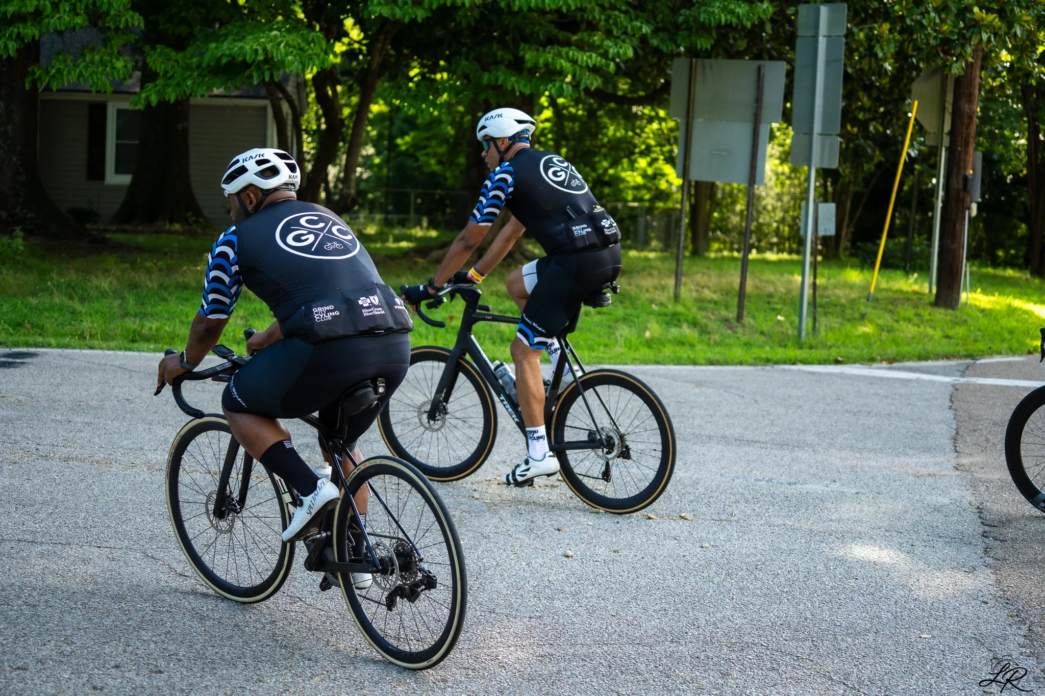 Two cyclists wearing helmets and matching cycling jerseys ride on a paved path surrounded by trees.