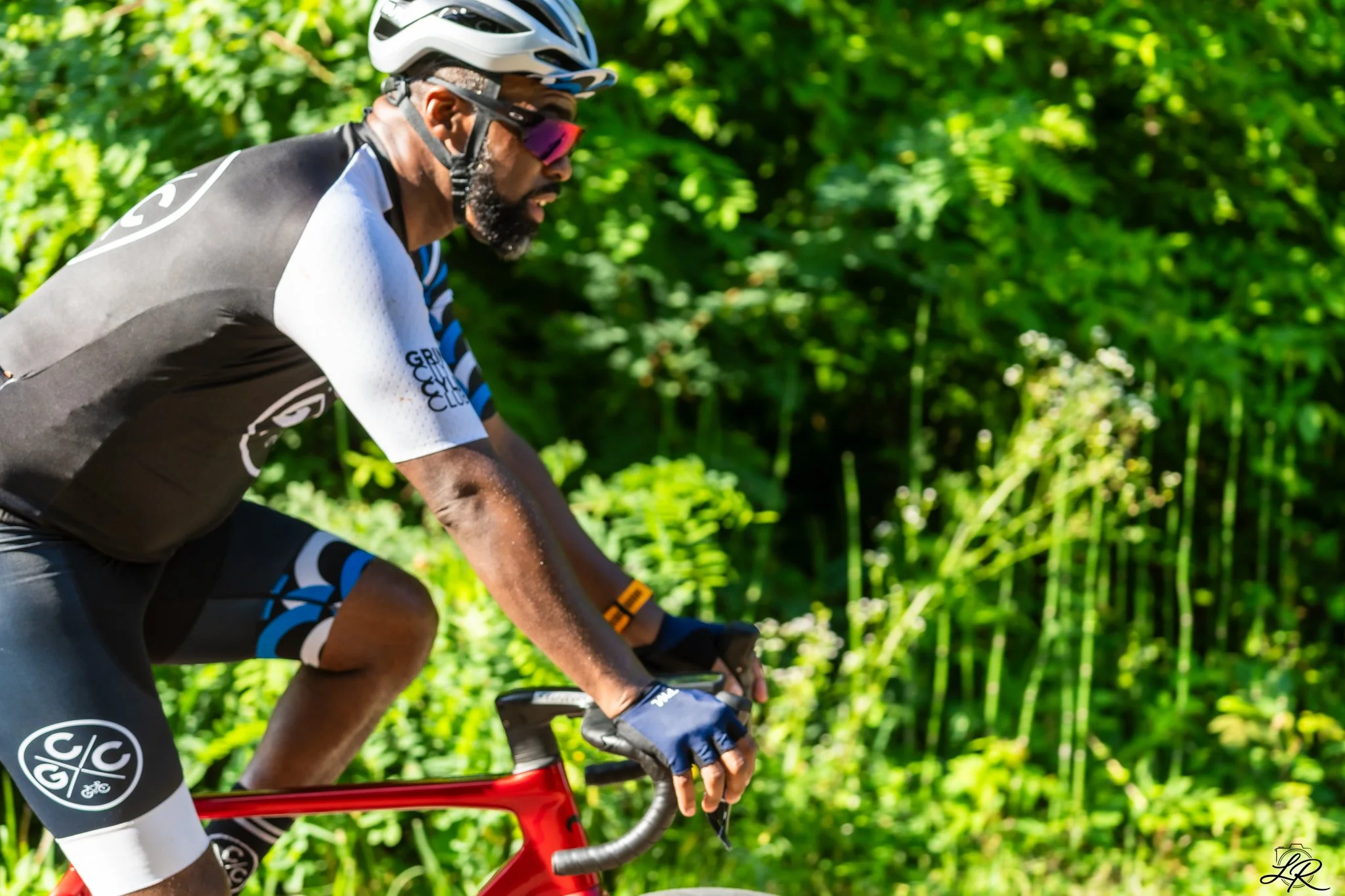 A man wearing a white cycling helmet, sunglasses, and a black and white cycling jersey riding a red bike through lush green surroundings.