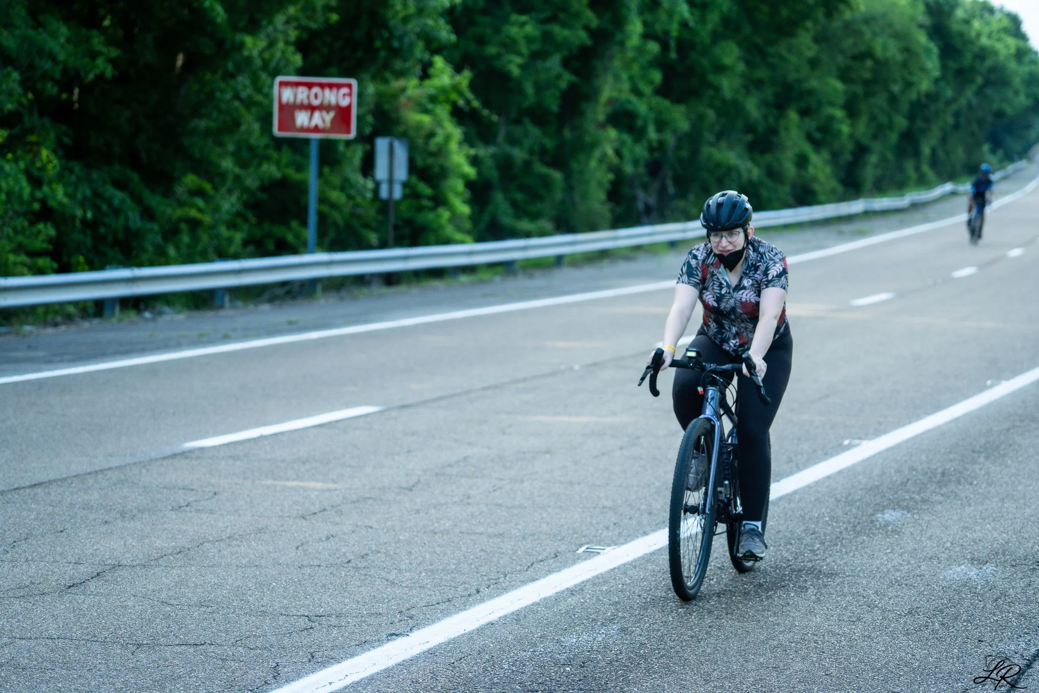 A person riding a bicycle on a road with a green forest background and a red sign that reads 'Wrong Way'.
