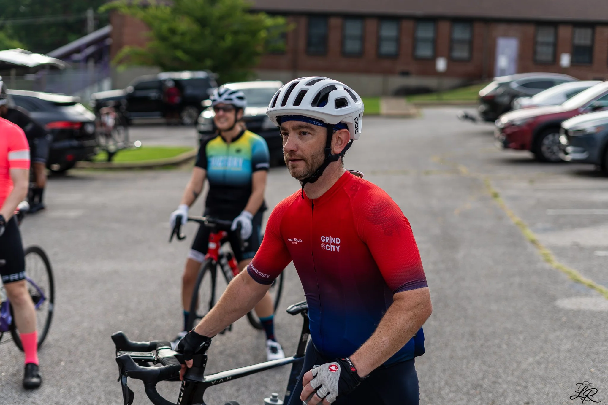 A group of cyclists gathered outdoors, with one man in red cycling attire and a white helmet in the foreground. Other cyclists are visible in the background, all wearing helmets and casual cycling gear. The setting appears to be a parking lot near a 