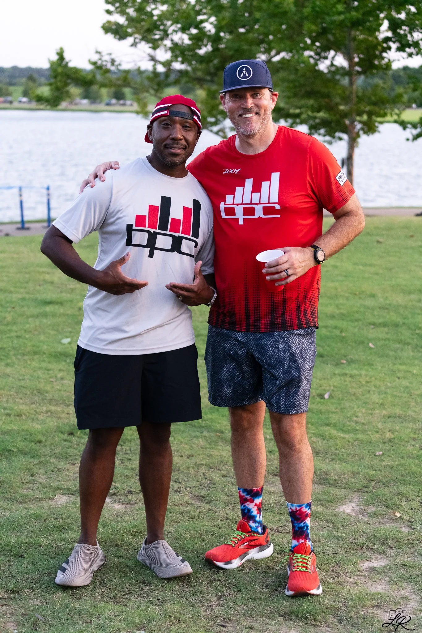 Two men standing outdoors on grass near a body of water, smiling at the camera. The man on the left is wearing a white sports shirt, black shorts, a red cap worn backwards, and sport shoes. The man on the right is wearing a red shirt, patterned short