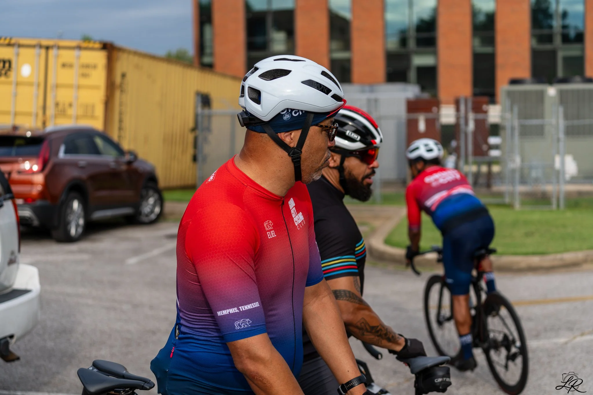 A group of cyclists sitting on their bikes at a rest stop, with a male cyclist in a red and blue jersey in the foreground wearing a white helmet and sunglasses. Other cyclists are seen behind him, also wearing helmets and cycling gear. There are cars