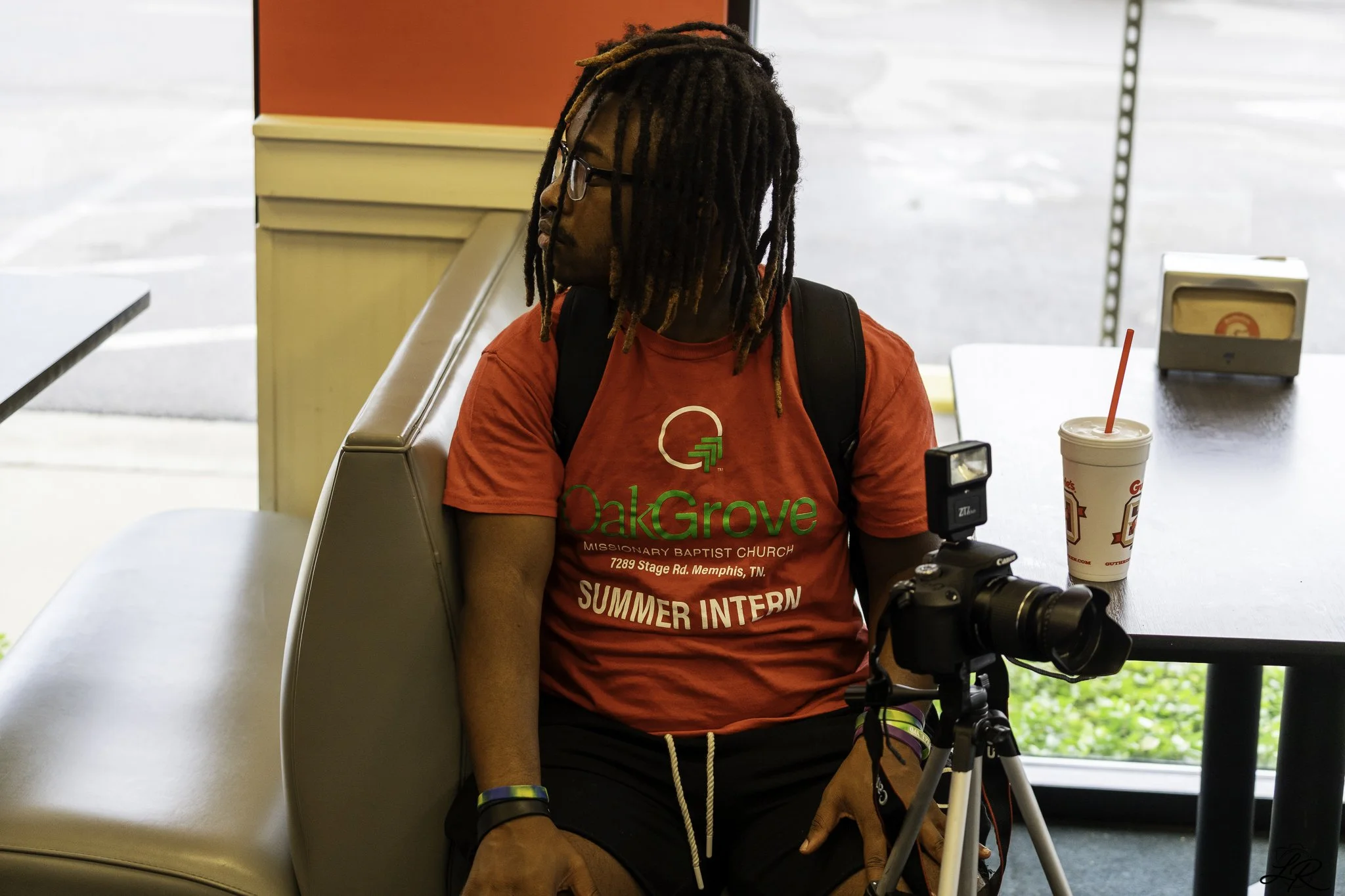 A young man with long dreadlocks and glasses sitting on a bench in a fast food restaurant, wearing a red T-shirt that says OakGrove Summer Intern, with a drink cup and a camera on the table beside him.