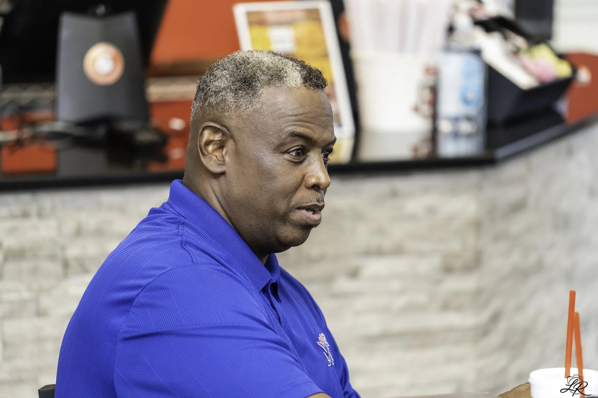 A middle-aged man with short gray hair wearing a blue polo shirt, sitting at a table in a fast-food restaurant, with a blurred background of food items and condiments.