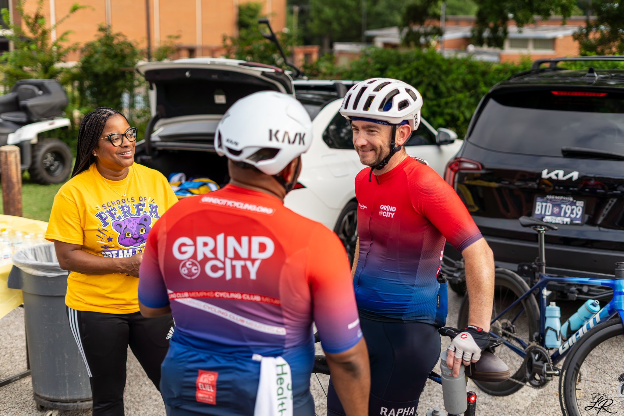 Two male cyclists wearing helmets and colorful cycling jerseys talking to a woman wearing glasses and a yellow T-shirt with a purple bear logo at outdoor event with cars in background.