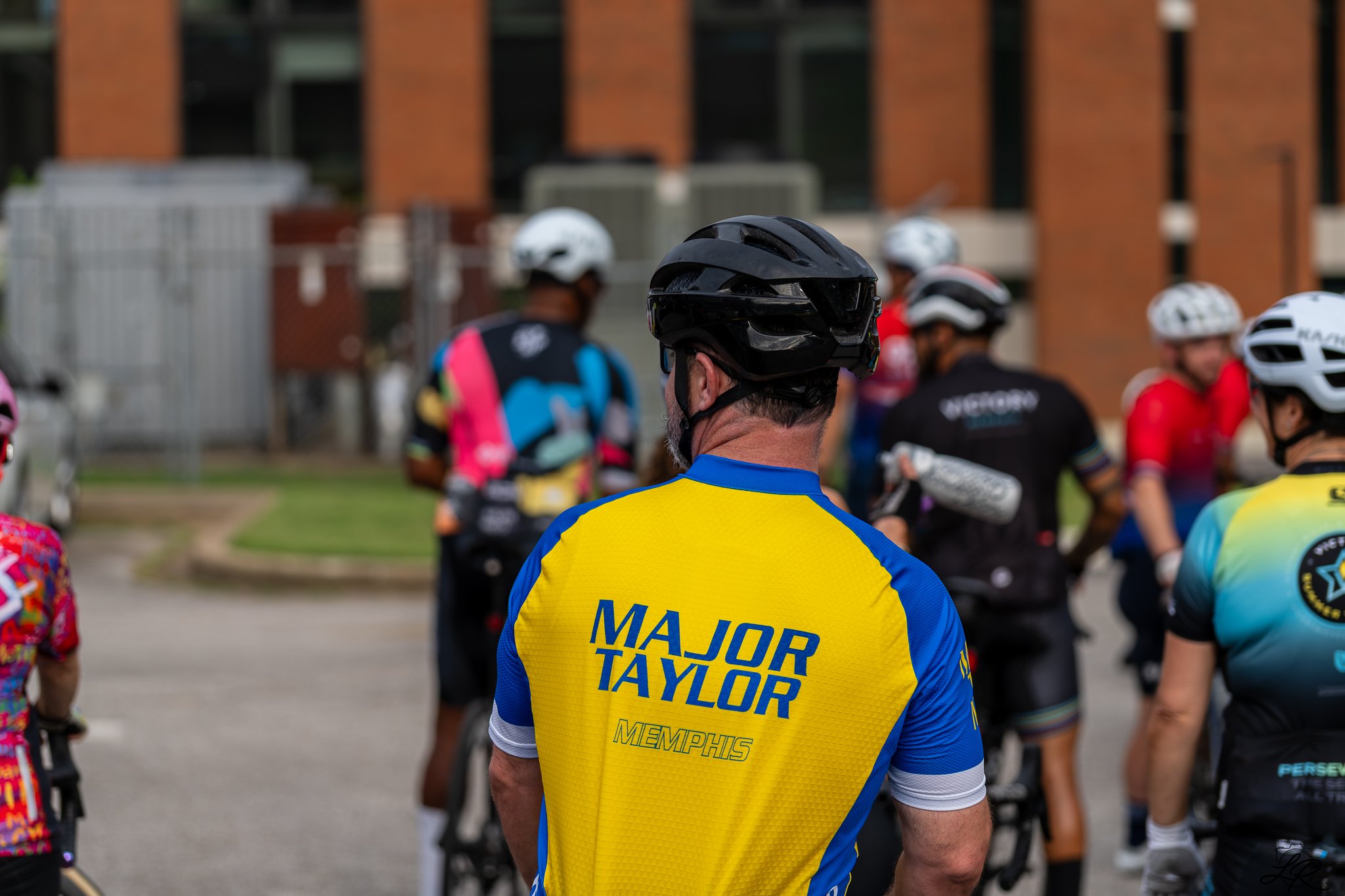A group of cyclists wearing helmets and colorful cycling jerseys gather together on a street with a brick building in the background. The focus is on a person wearing a yellow and blue shirt with the name 'Major Taylor Memphis' on the back.