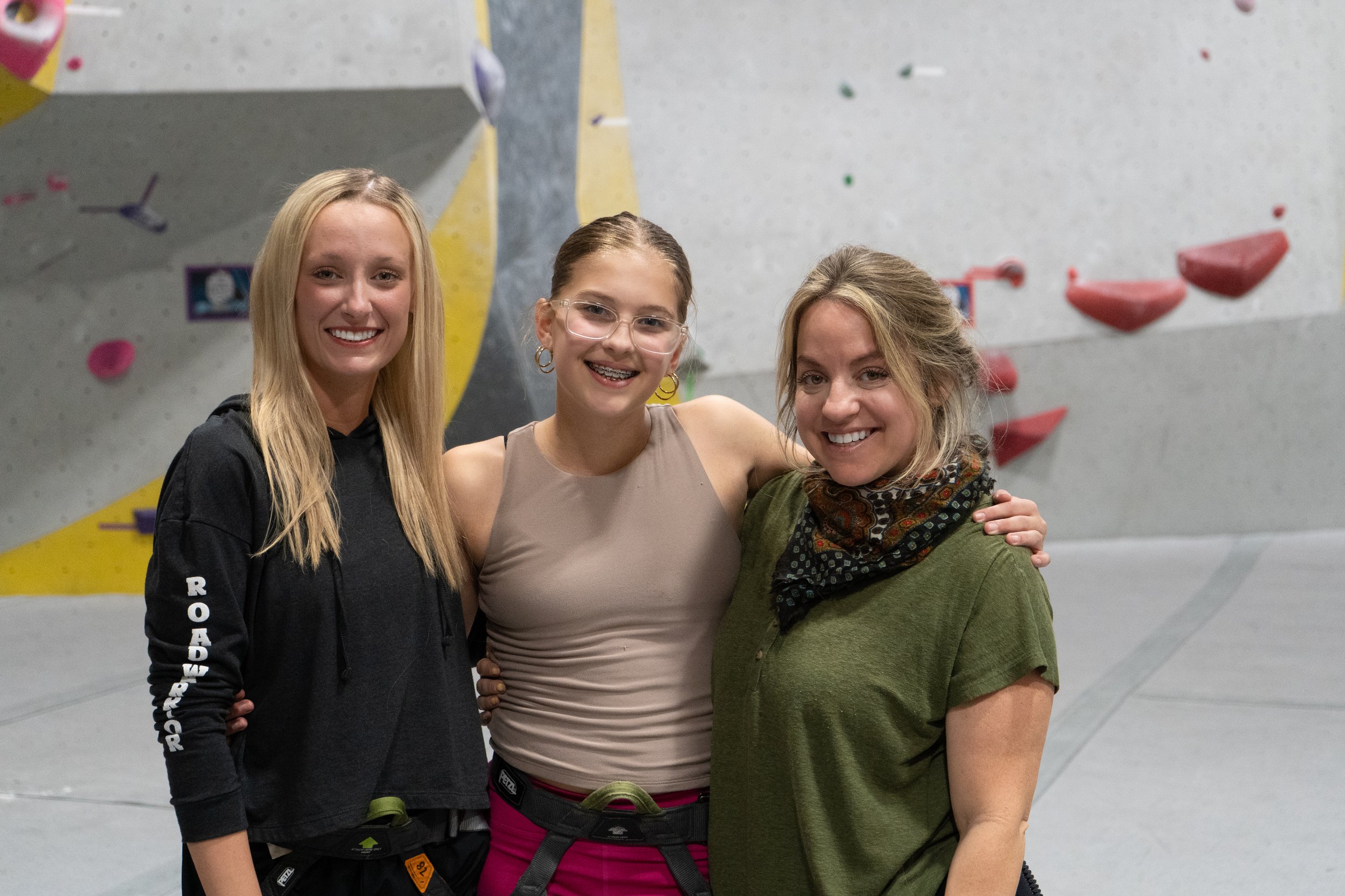 Three smiling women at an indoor climbing gym, standing in front of a gray climbing wall with red, yellow, and purple holds.