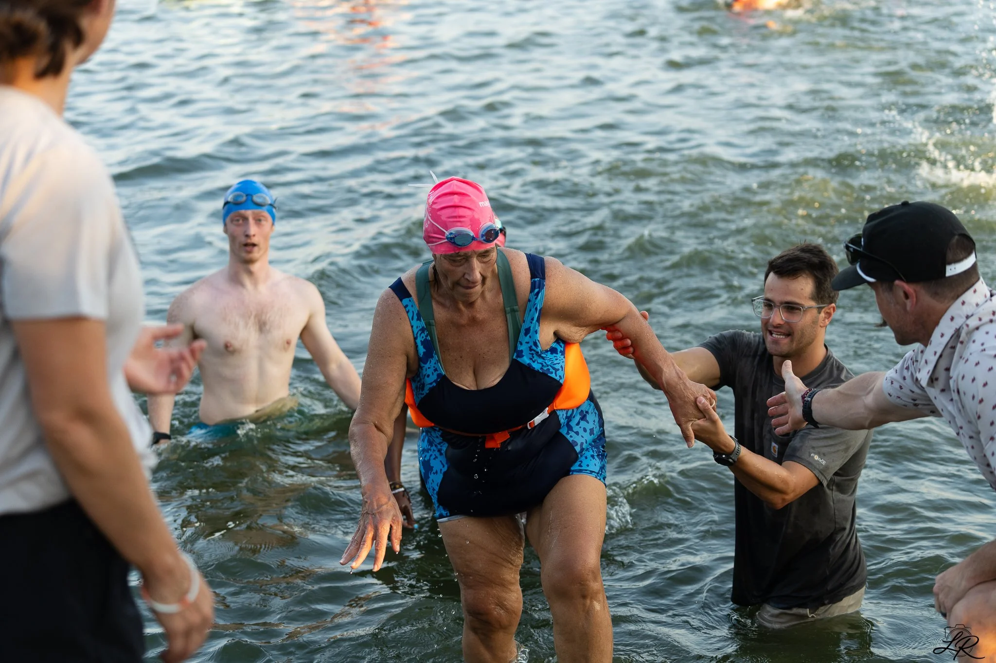 A woman in a pink swimming cap and goggles is being helped out of the water by several people at a swimming event near a body of water.