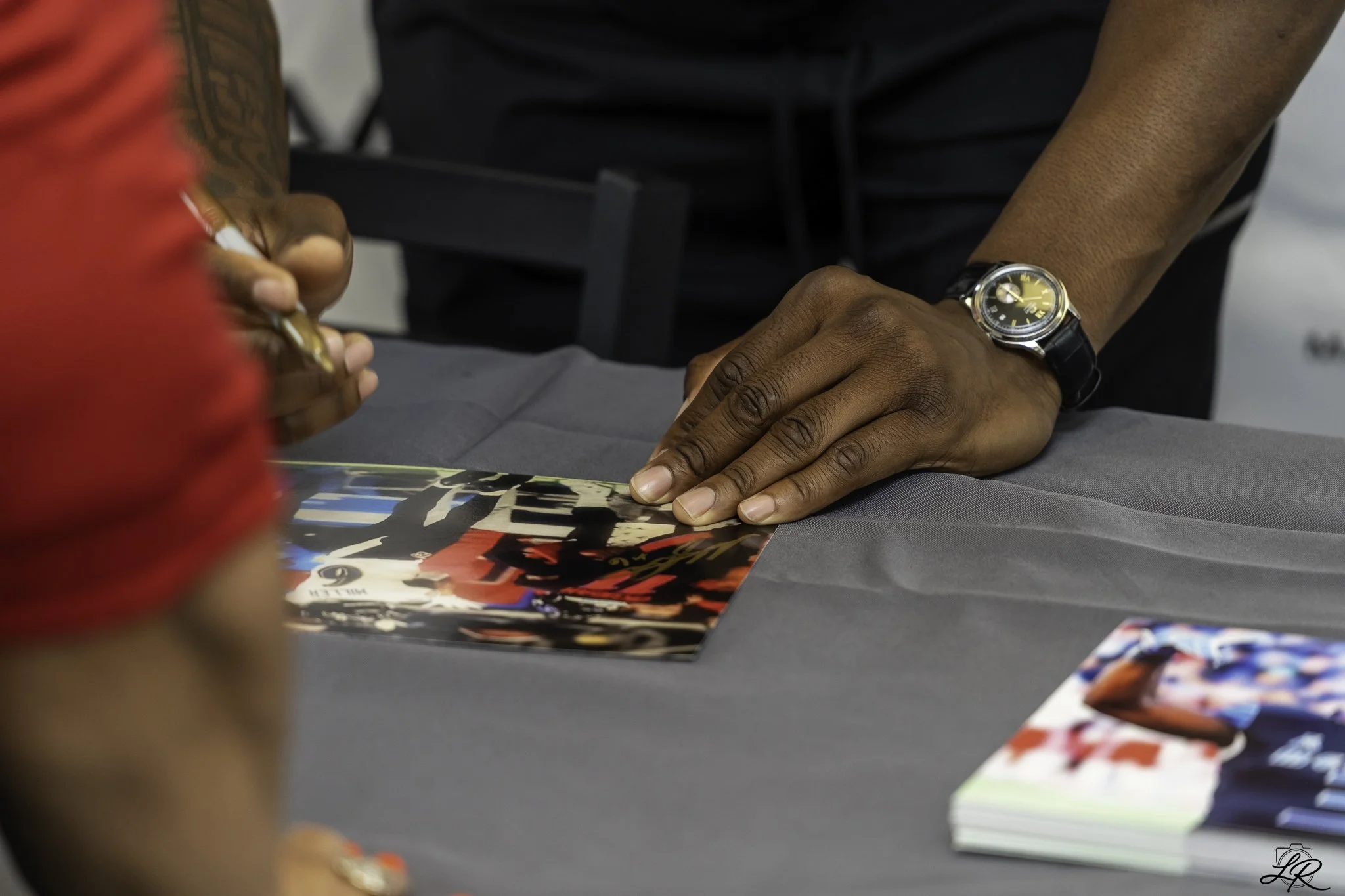 Close-up of a person's hand wearing a wristwatch, resting on a table with colorful magazine or brochure and another person's hand holding a pen. The background is blurred, showing more magazines or brochures on the table.