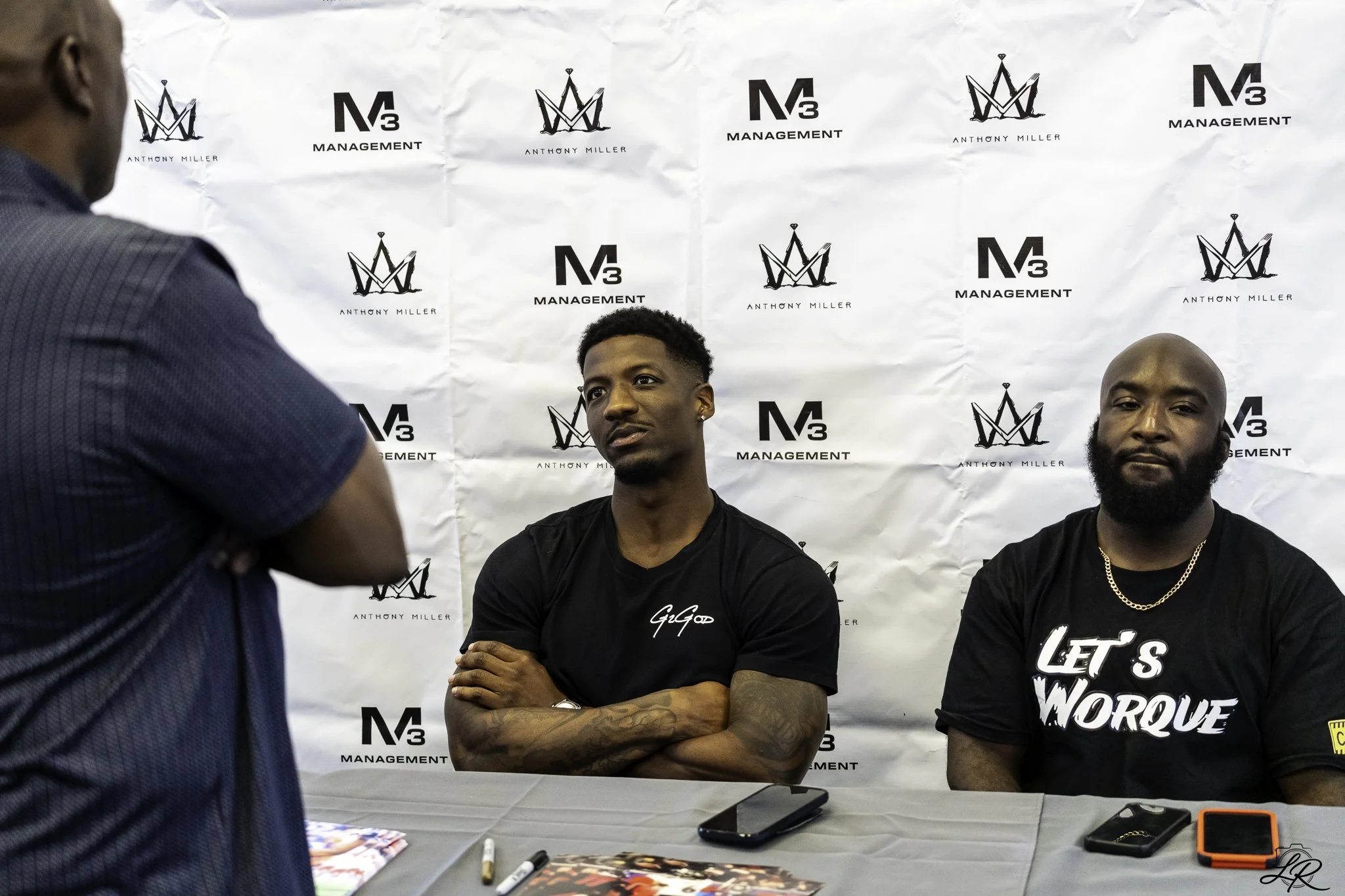 Two men seated behind a table with merchandise, facing a man in a blue shirt, at a promotional event with a backdrop that has logos and text reading 'Anthony Miller' and 'Management.'