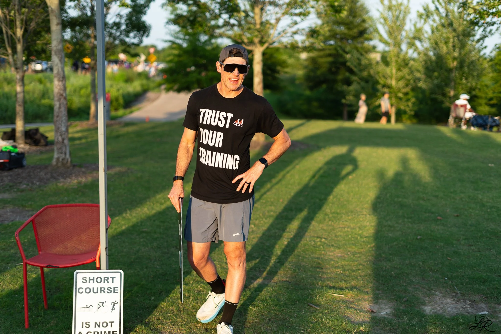 A man running on a short course with a sign that reads 'SHORT COURSE IS NOT A CRIME.' He is wearing sunglasses, a black cap backward, a black t-shirt with 'TRUST YOUR TRAINING' printed on it, shorts, and running shoes. The background shows trees, a p