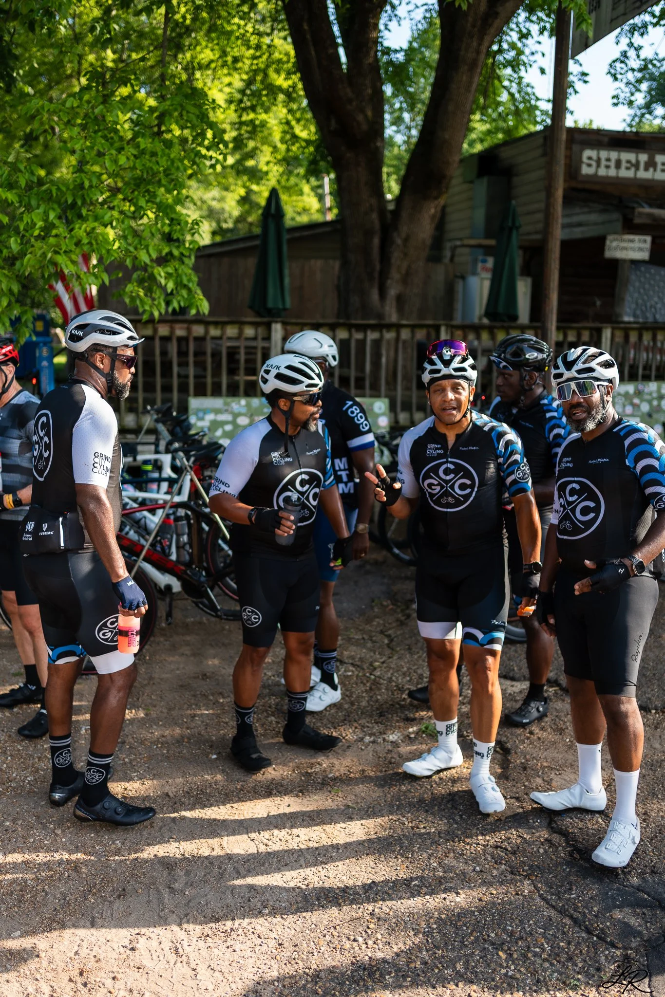 A group of cyclists wearing matching black and blue cycling jerseys and helmets stands together outdoors, engaged in conversation. They are near bicycles, with some holding water bottles. The background features green trees, a wooden fence, and a bui