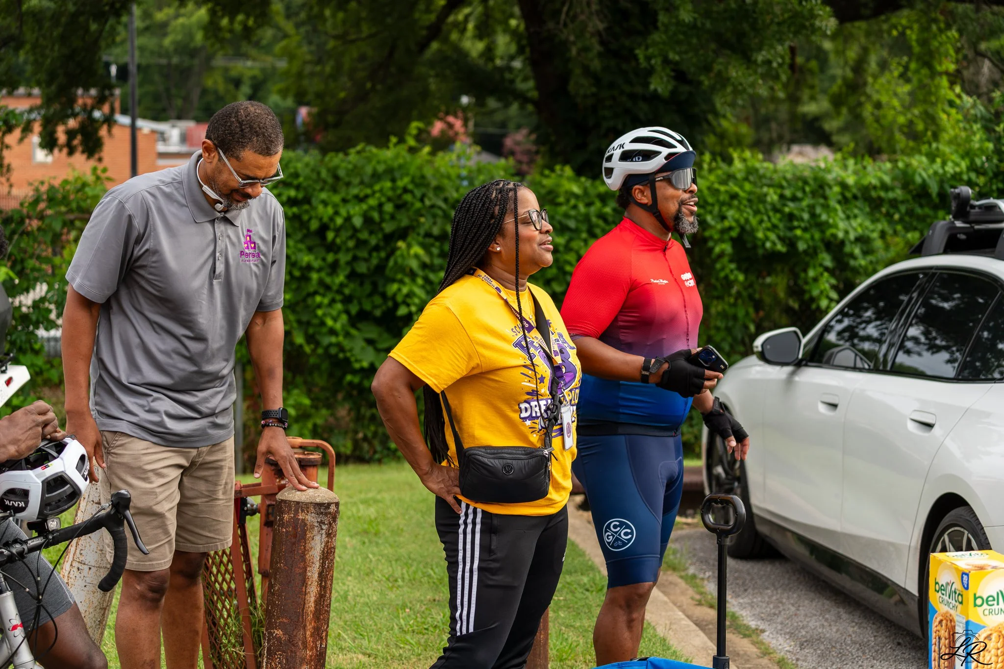 Three adults standing outdoors in front of a green hedge, one with a bicycle helmet, one with a yellow T-shirt, and one with sunglasses and cycling attire, participating in an event in a park.