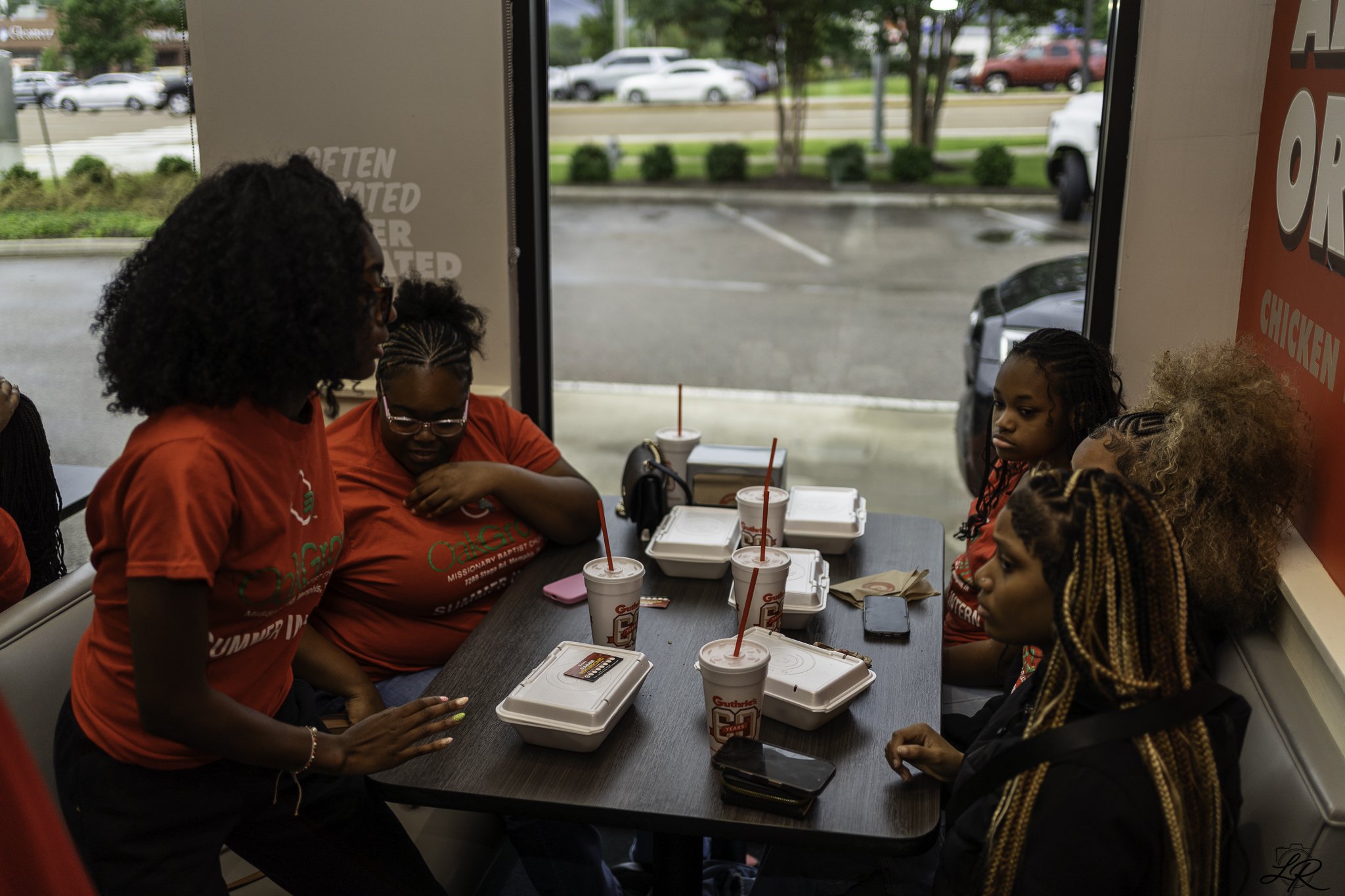 A group of women and girls sitting around a table inside a fast food restaurant. They are wearing matching red shirts, and there are several food containers and drinks on the table. The scene is viewed through a window with a parking lot and trees ou