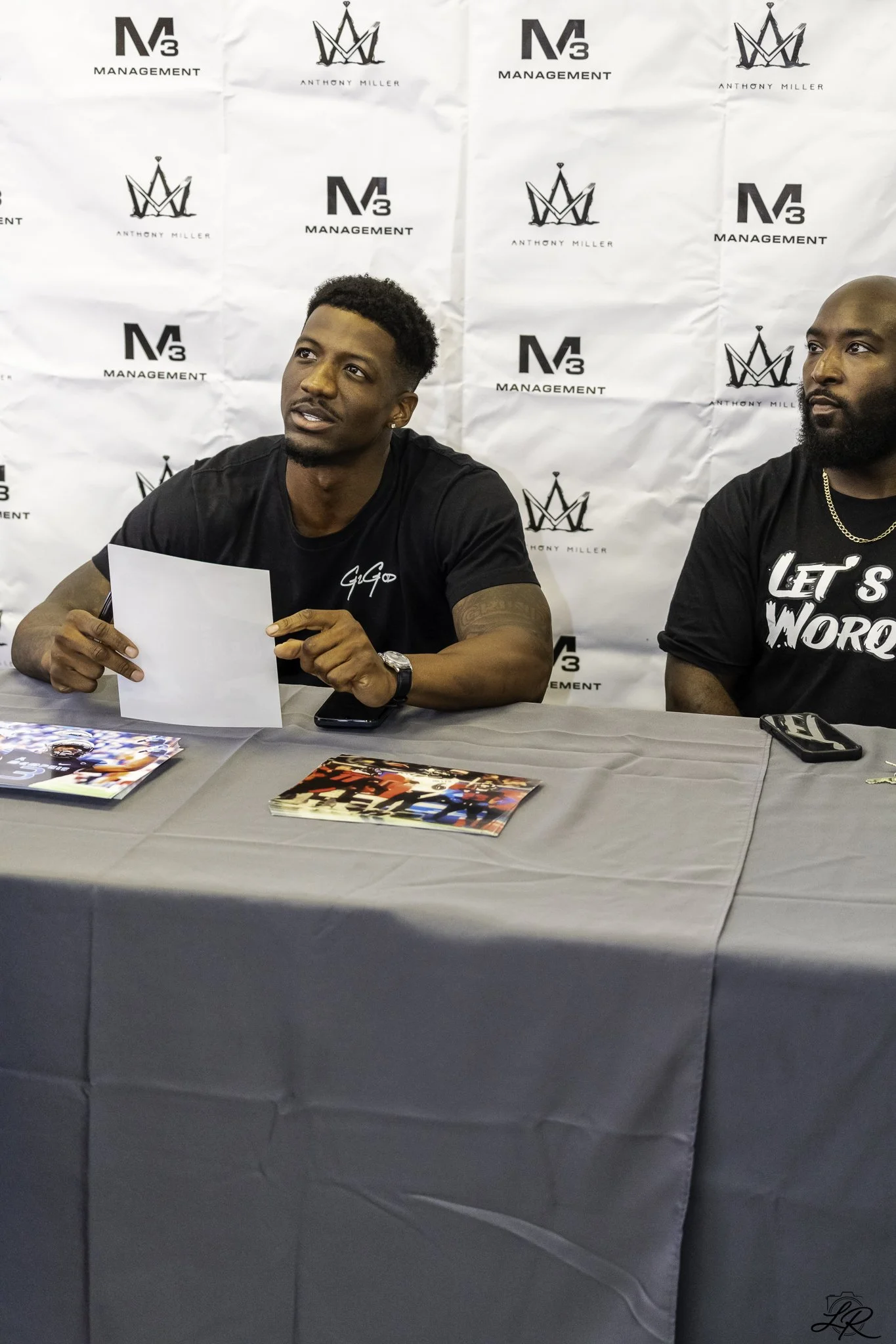 Two men sit at a table during a promotional event or signing. The man on the left holds a piece of paper, while the man on the right wears a black shirt with text. The backdrop has logos for 'M3 Management' and 'Anthony Miller'. On the table are maga