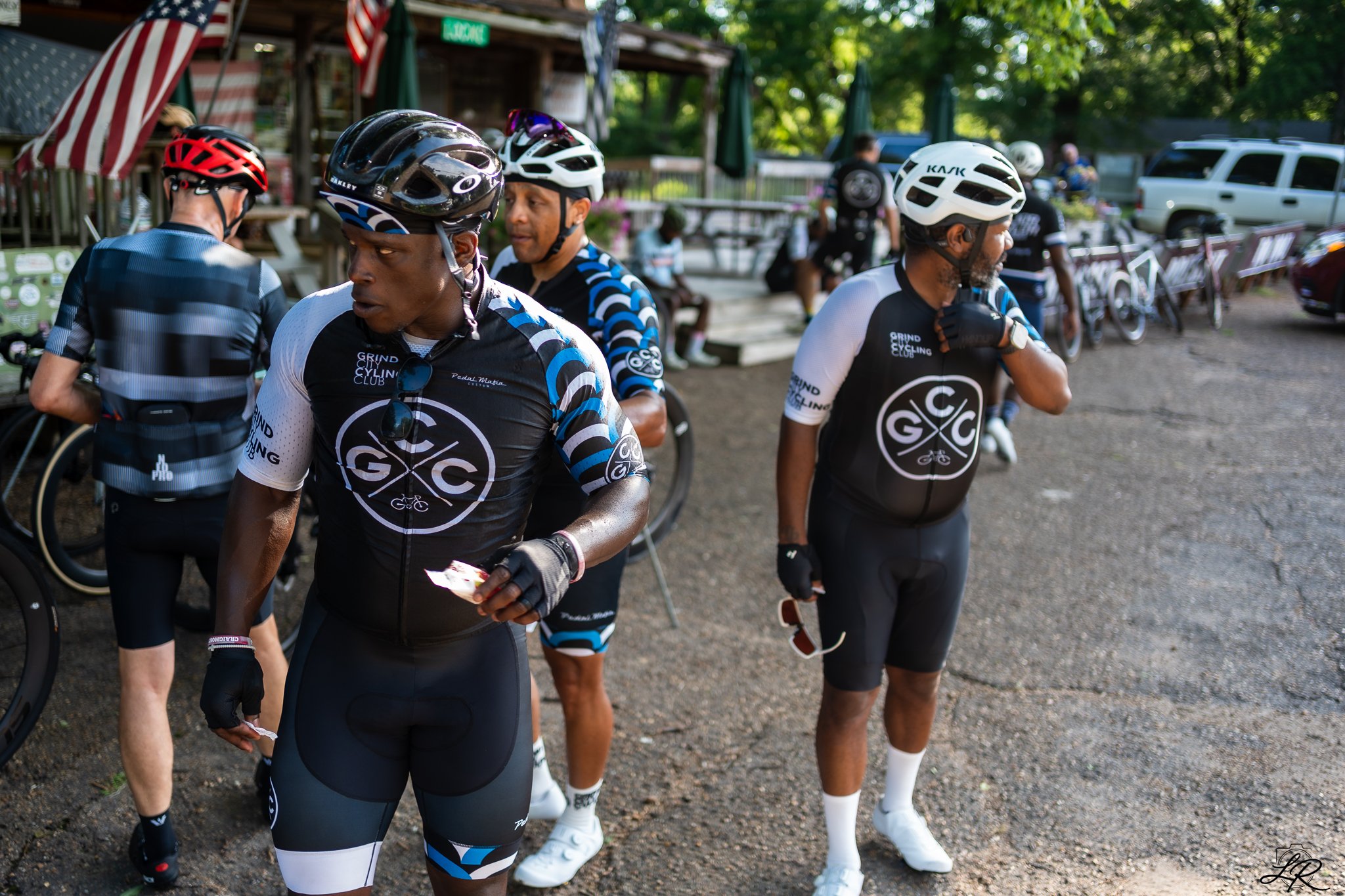 Group of cyclists in cycling gear and helmets standing outdoors near bikes, with a wooden building and trees in the background.