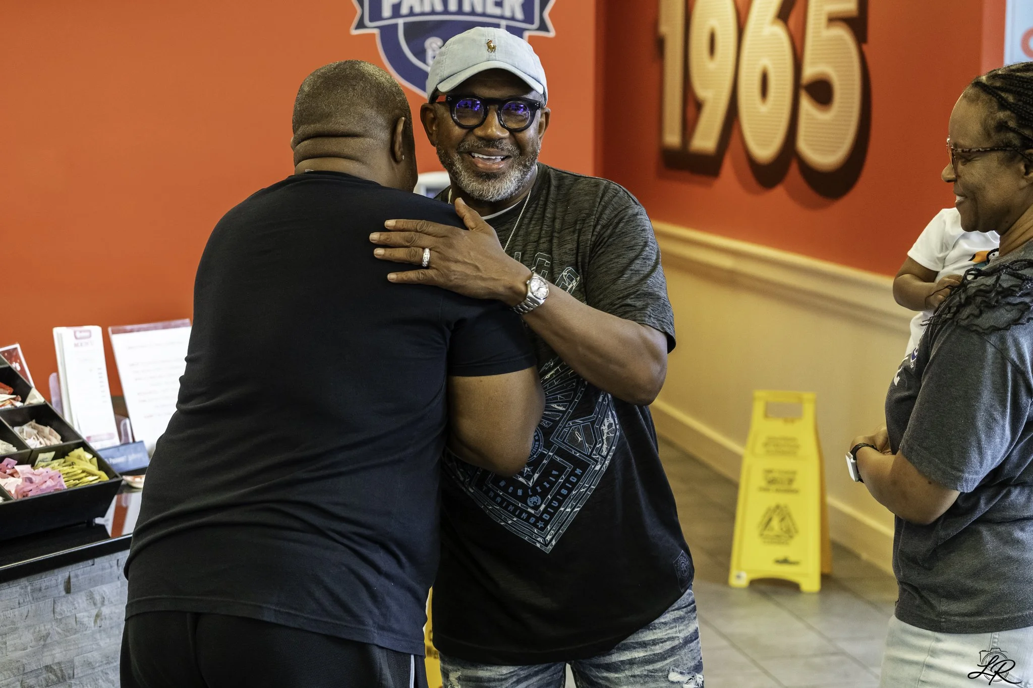 Three people smiling and engaging in a friendly hug at a casual indoor setting, with a red wall featuring a partially visible logo and a large 195 sign in the background. One man is wearing sunglasses, a light blue cap, and a black T-shirt. A woman w
