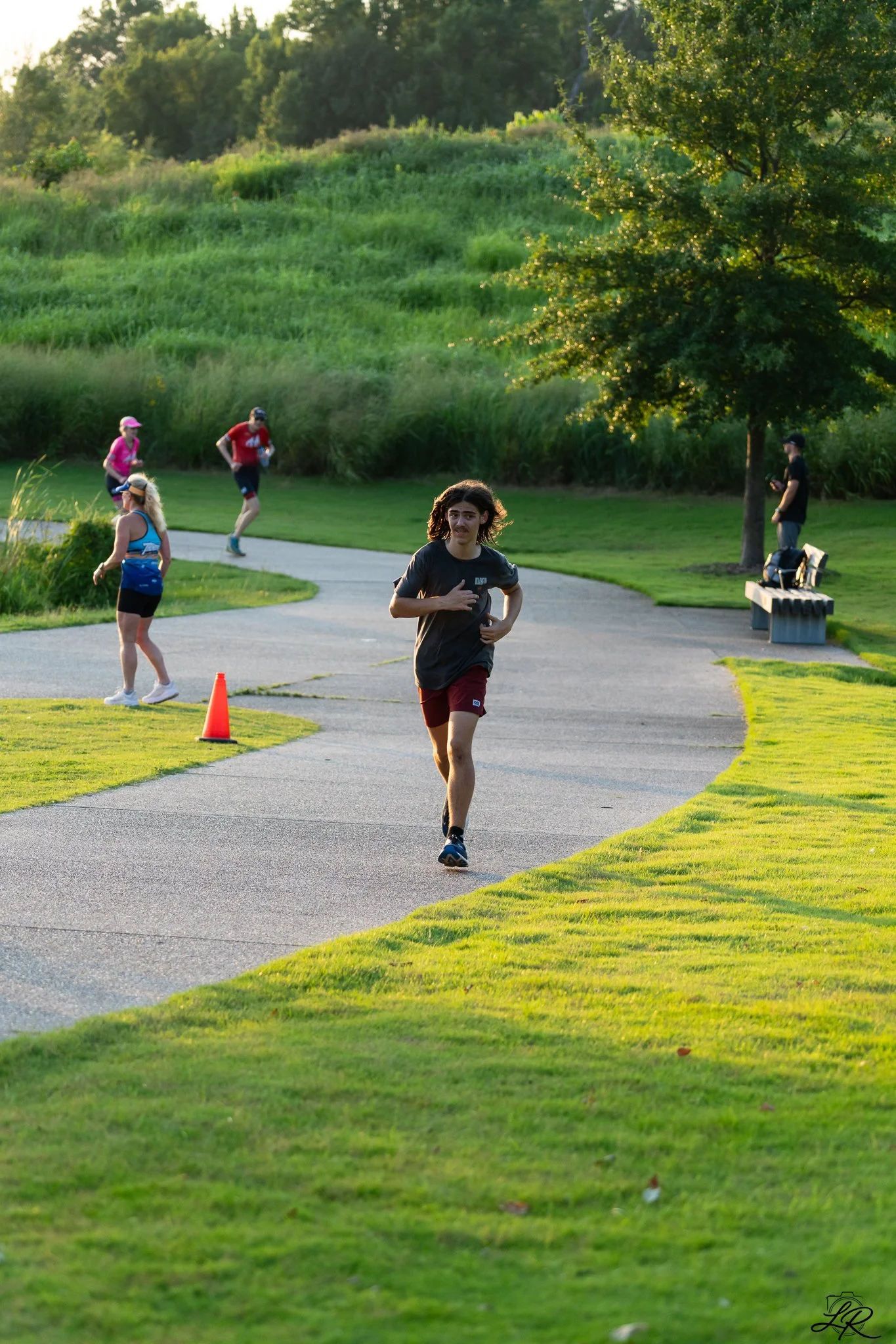 A group of people jogging and walking along a curved paved path in a lush green park during late afternoon or early evening sunlight, with trees and grassy hills in the background.