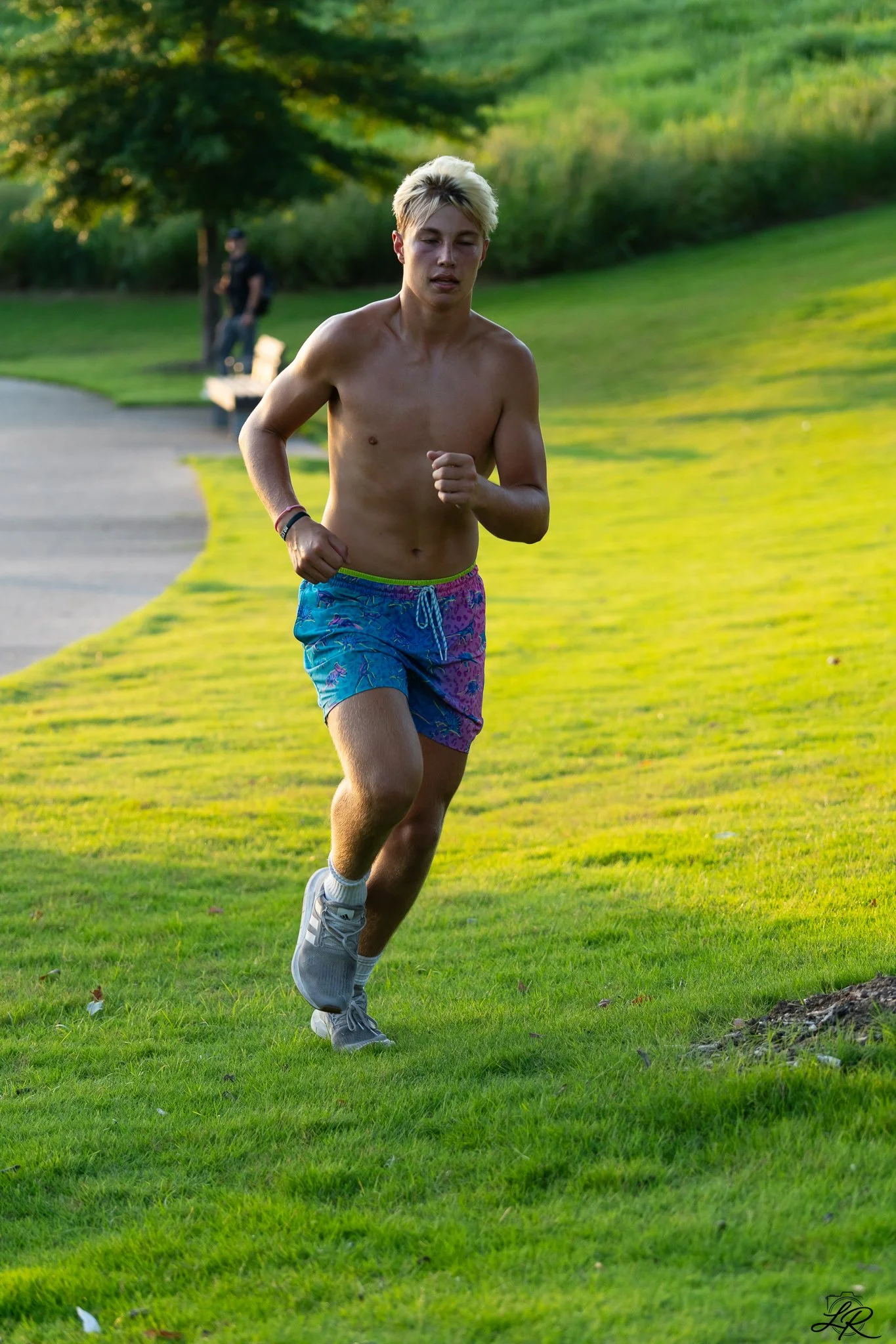 A shirtless young man with blonde hair running on a grassy park path during late afternoon or early evening, with another person sitting on a bench in the background.