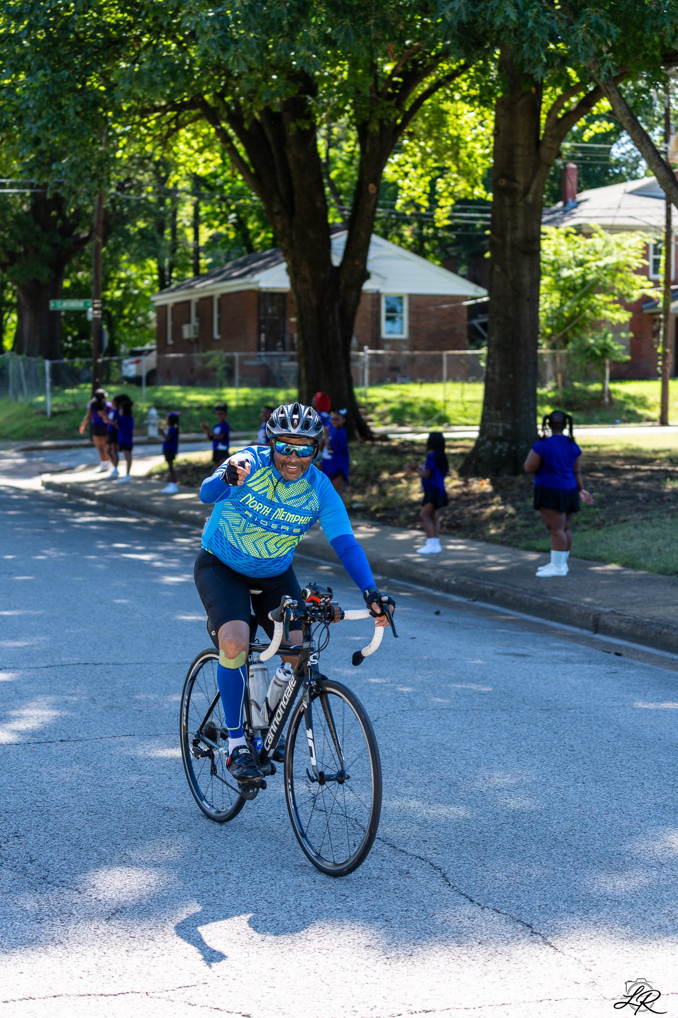 A man wearing a blue cycling jersey, black shorts, blue knee-high socks, a helmet, and sunglasses riding a gray Cannondale road bike on a street with a group of children in blue uniforms and a large tree with green leaves in the background.