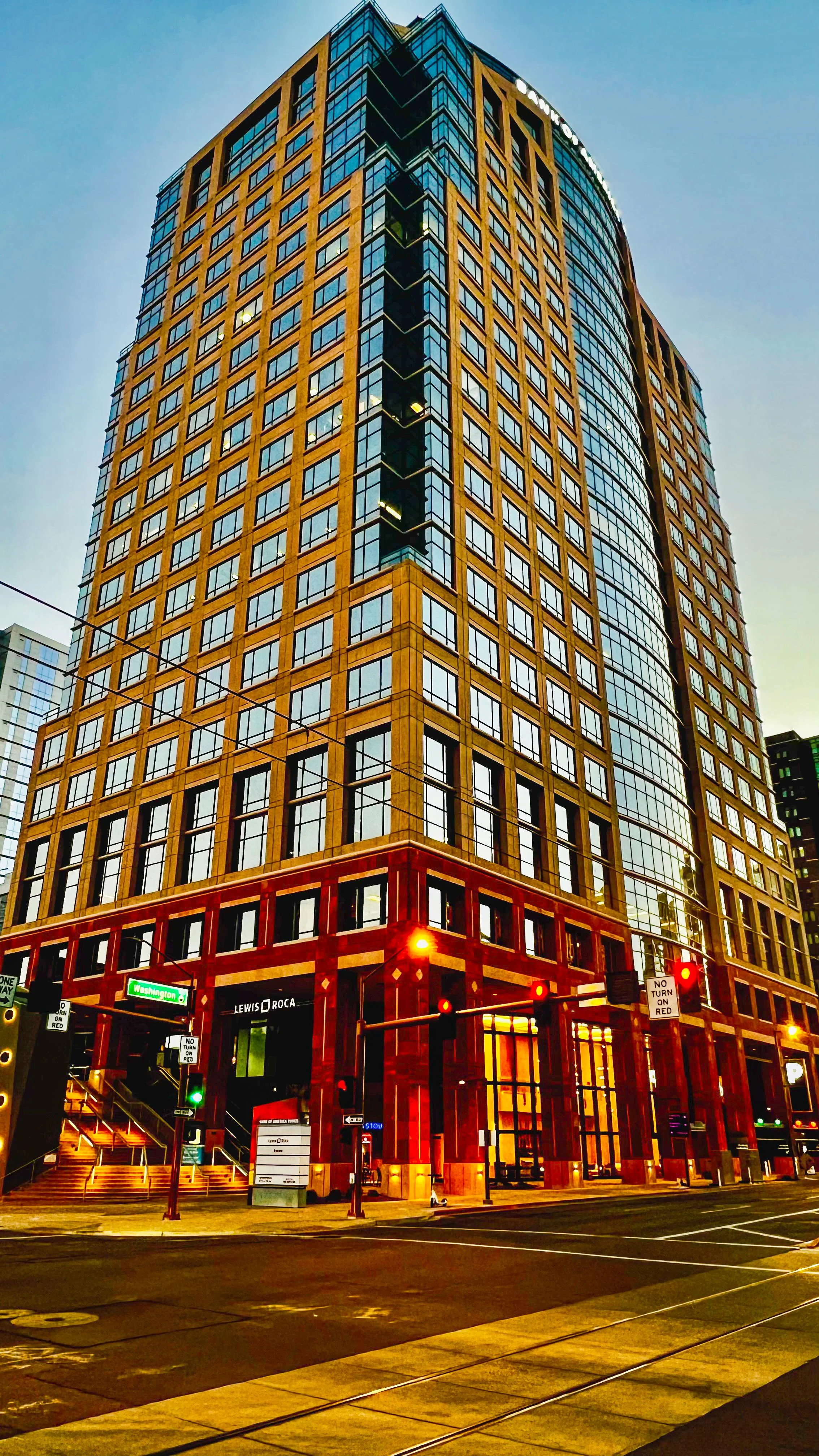 A tall modern building with a blue glass and tan facade, located at the corner of an intersection during dusk or dawn, with streetlights and traffic signals illuminated.