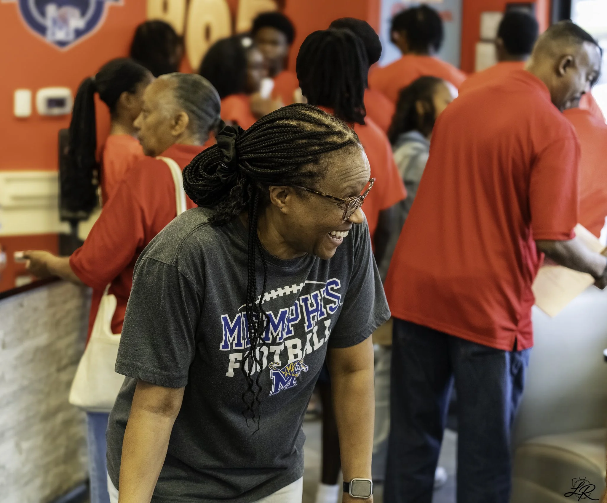A diverse group of people standing in a line, wearing Memphis football shirts, in an indoor setting with red walls and Memphis Tigers decorations.