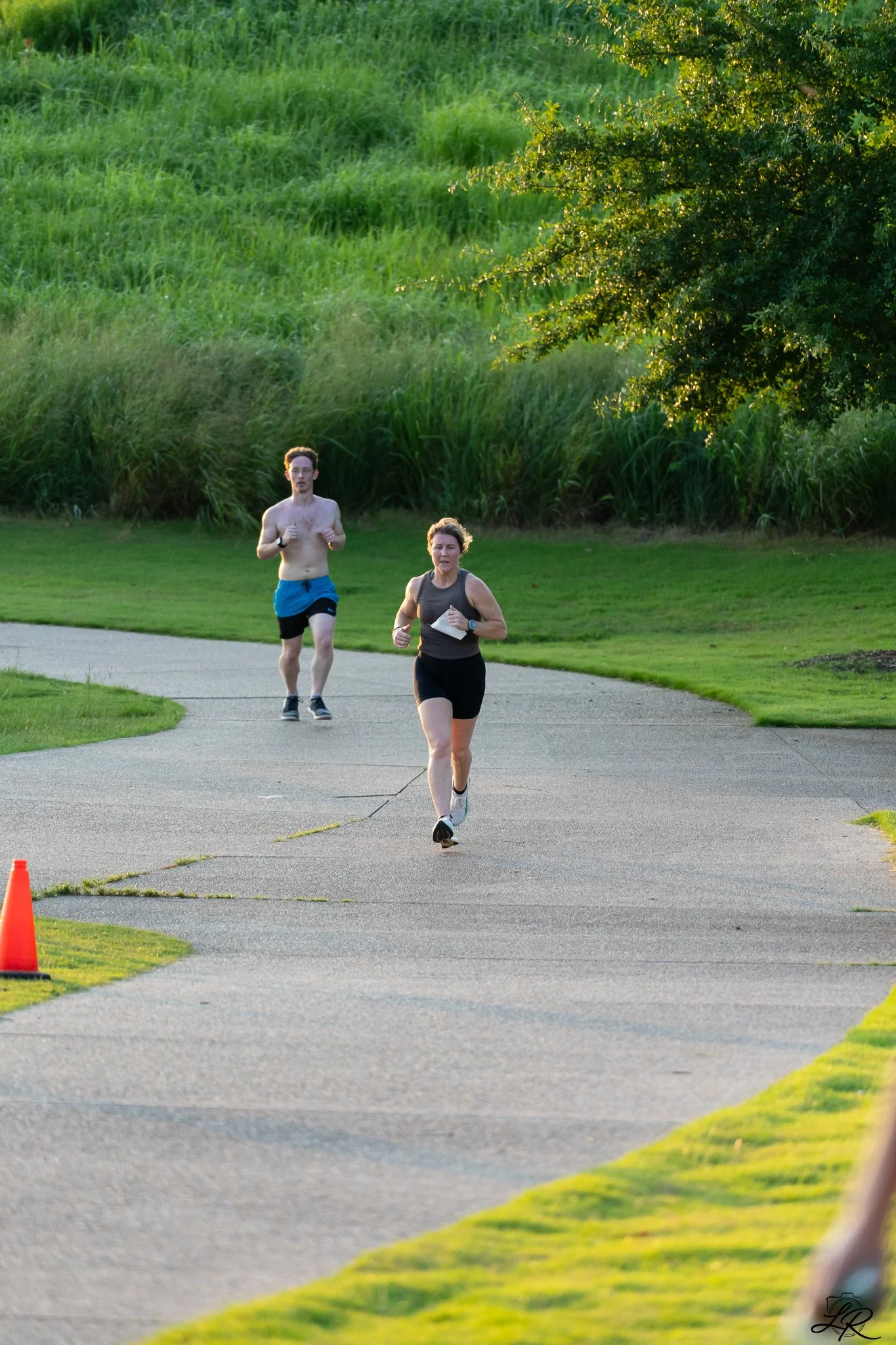 Two runners, a man without a shirt and a woman in a black tank top, running on a paved trail in a park with green grass and trees, during what appears to be early evening.