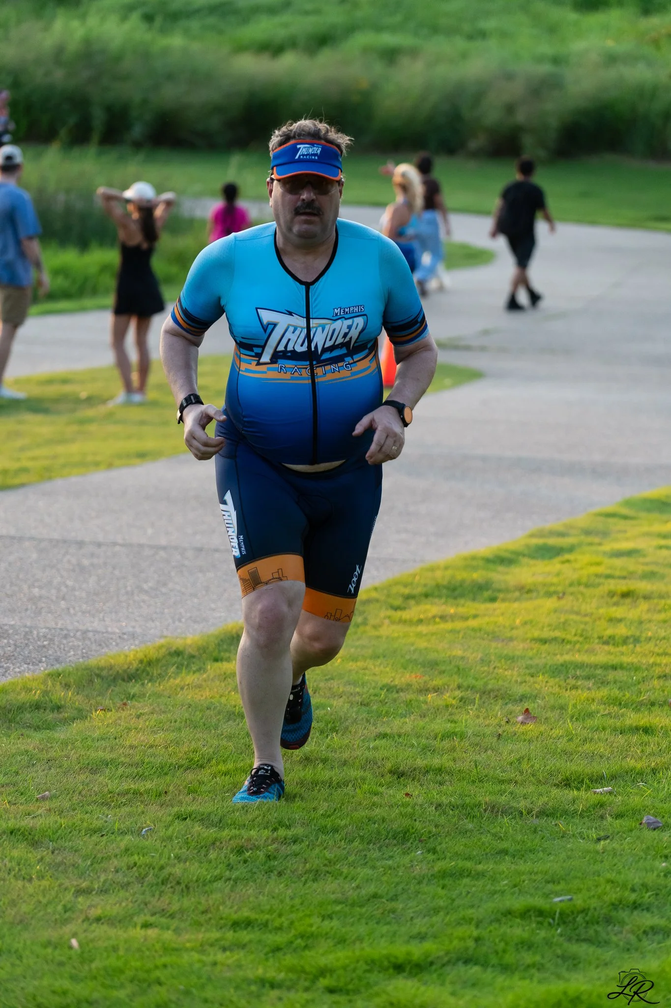 A man wearing a blue sports jersey with the logo 'Memphis Thunder Racing' runs on grass near a paved walkway, with several people walking in the background, in a park-like setting.