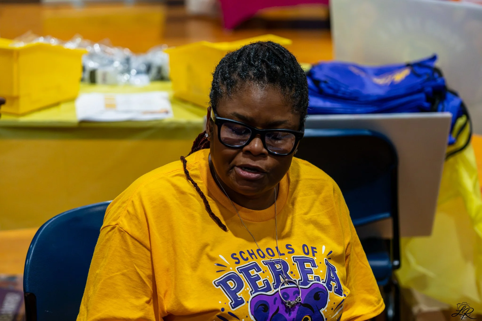 A woman with dark hair and glasses wearing a yellow shirt that says 'Schools of' and 'Peace' in large letters, sitting in a chair at an event with yellow and blue supplies in the background.