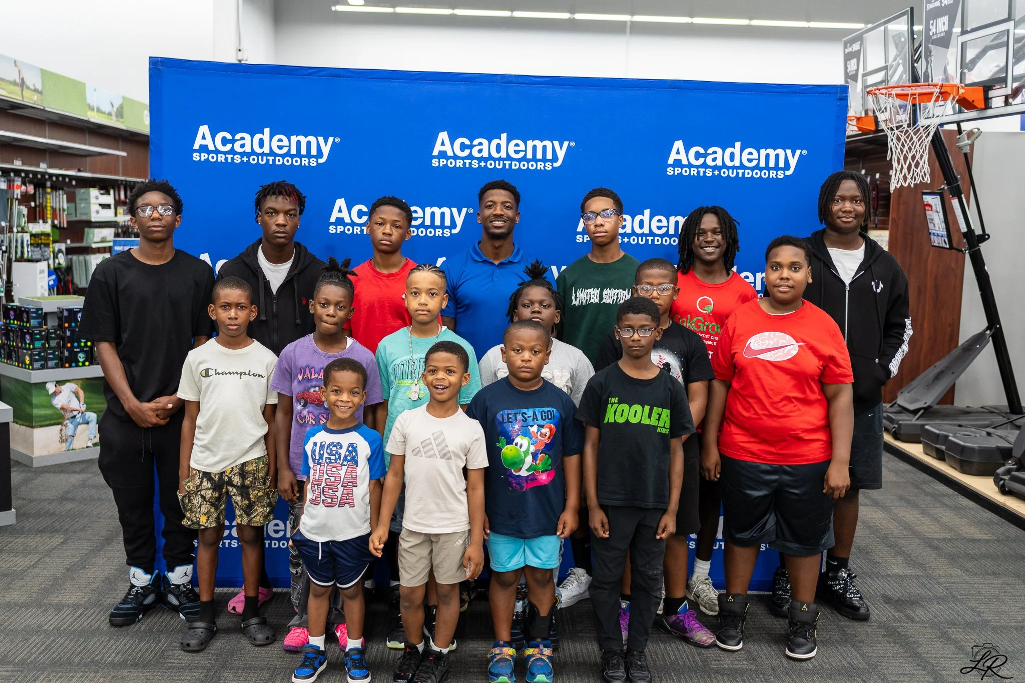 A group of children and teenagers posing for a photo in front of a blue backdrop with the 'Academy Sports + Outdoors' logo at an indoor sports store. Some children are smiling, others have neutral expressions. The background shows shelves with produc