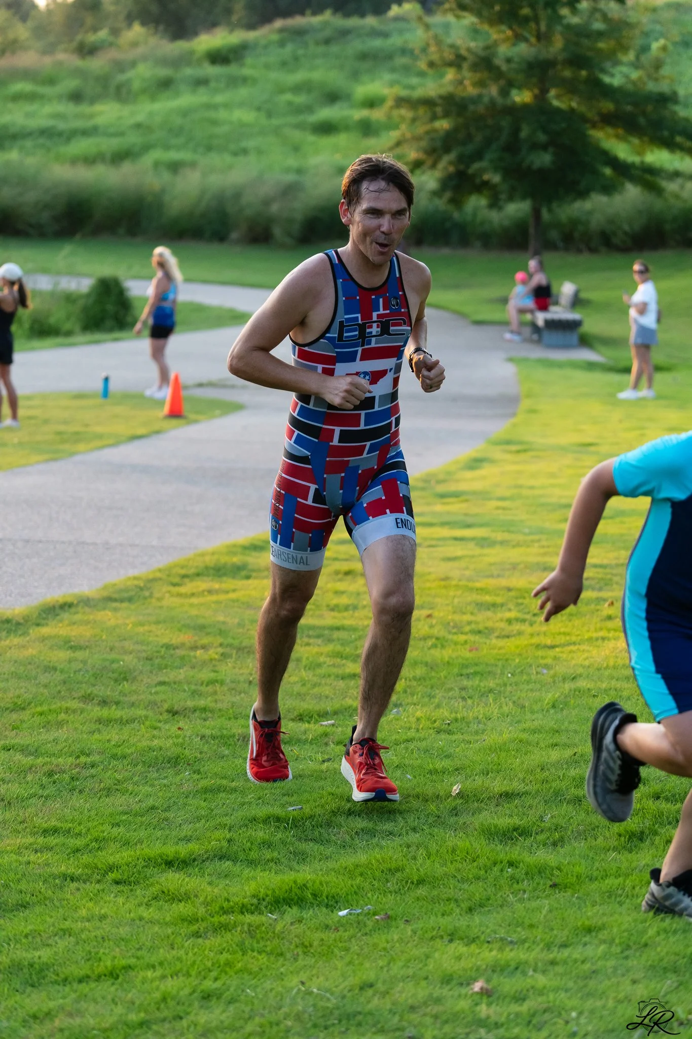 A man wearing a colorful patterned triathlon suit and red running shoes running outdoors on a grassy area during a race, with several spectators watching and taking photos in the background.