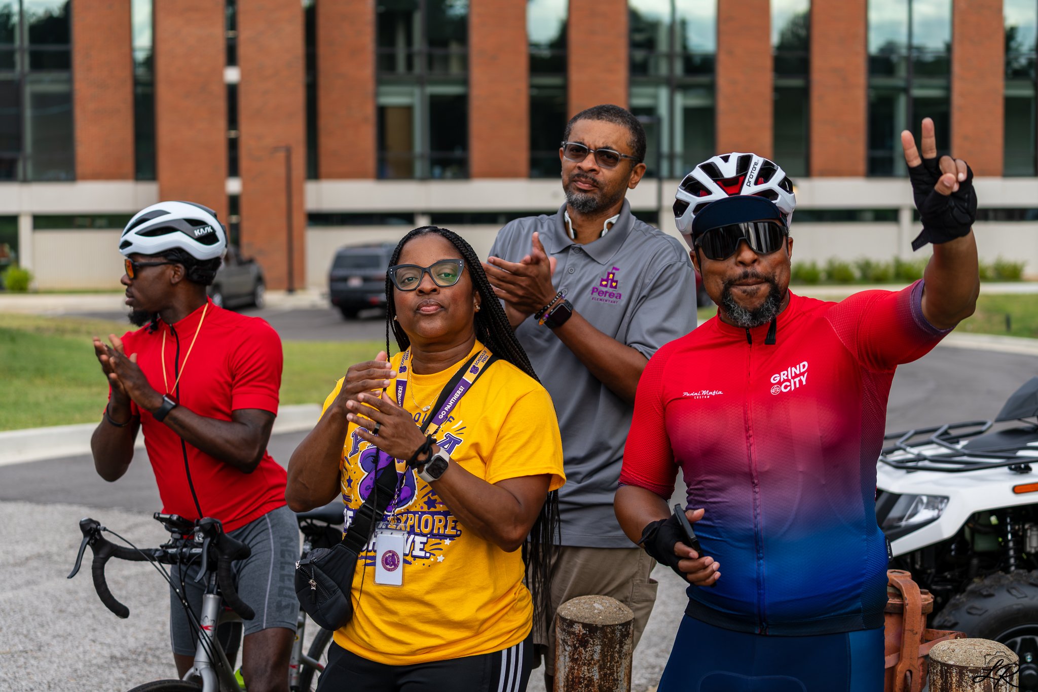 A diverse group of four adults, three men and one woman, standing outside in front of a brick building, dressed in athletic clothing with helmets and bikes, participating in an outdoor activity or event.
