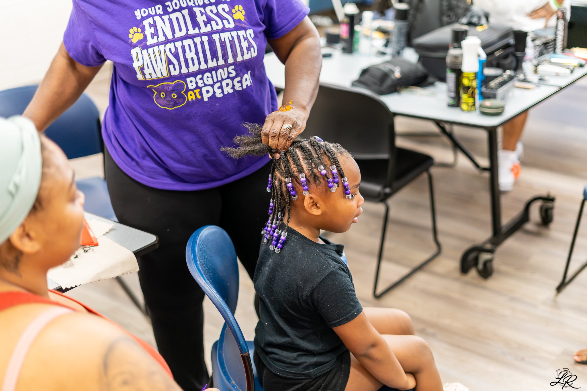 A woman with gray hair sitting in a classroom, having her hair braided by another woman wearing a purple T-shirt that says "Your journey to endless paw-sibilities begins at P'Rea". Other students are visible, along with tables and various hair care s