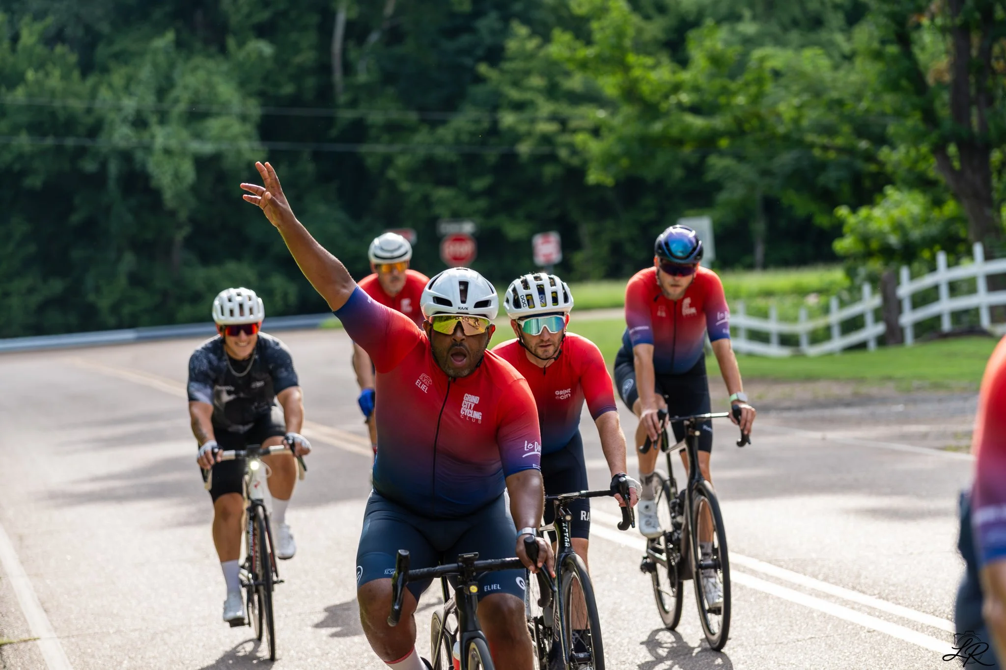 Group of cyclists riding on a road surrounded by trees, with one man in the front raising his hand.