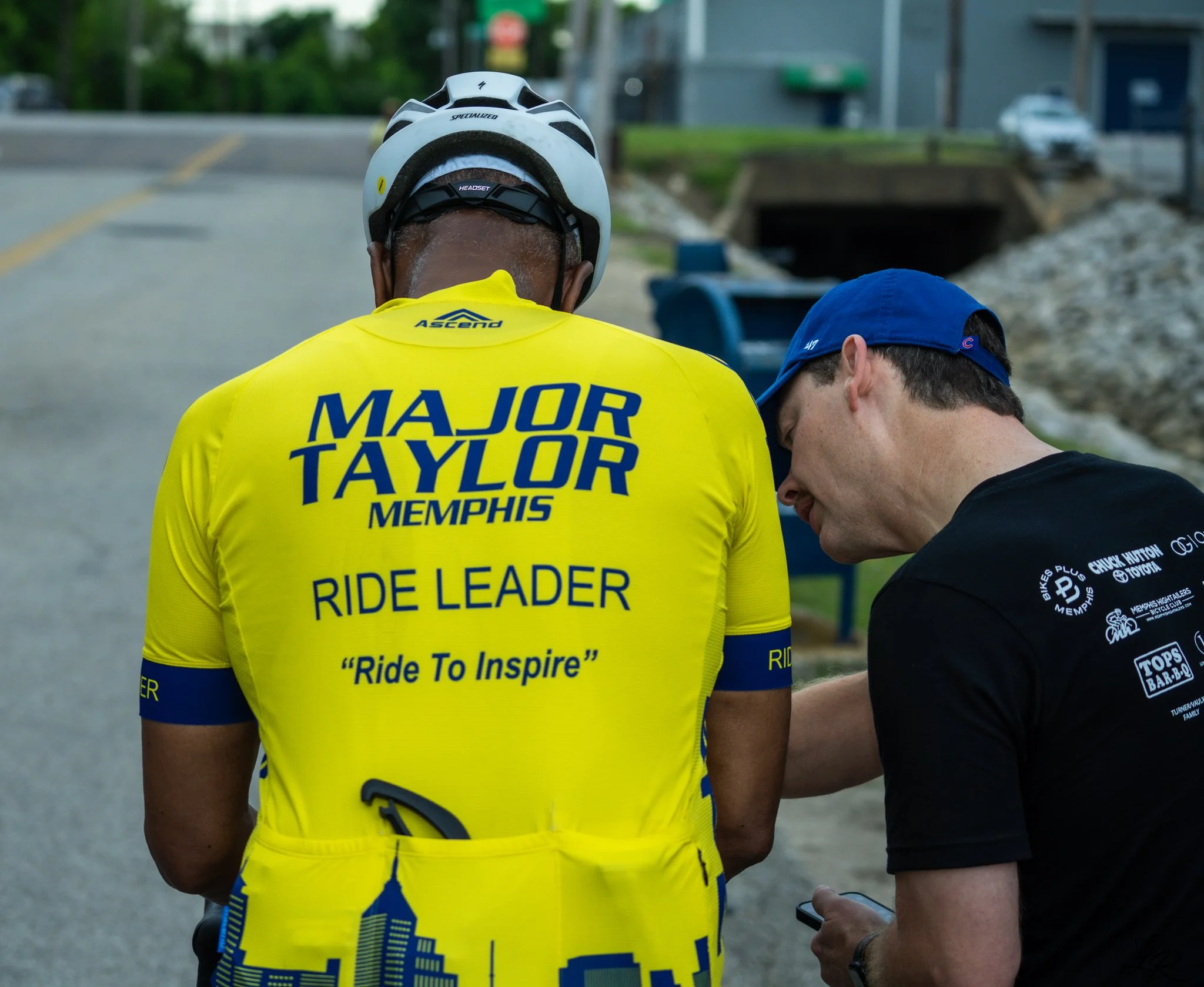 Person wearing a yellow cycling jersey with blue text that reads 'Major Taylor Memphis Ride Leader.' Person is also wearing a white helmet with black accents.