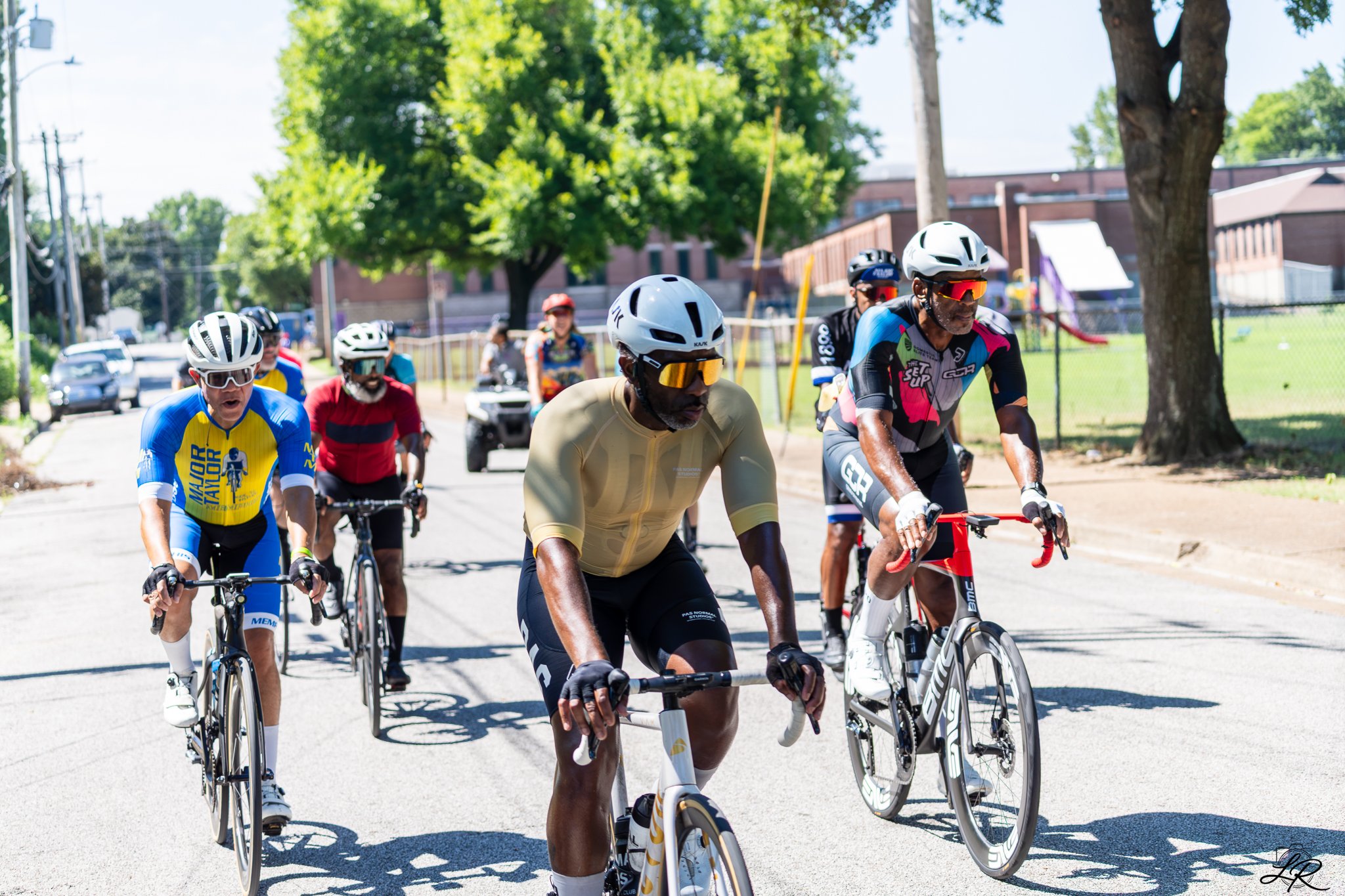 Group of cyclists riding on a sunny street with trees and buildings in the background.
