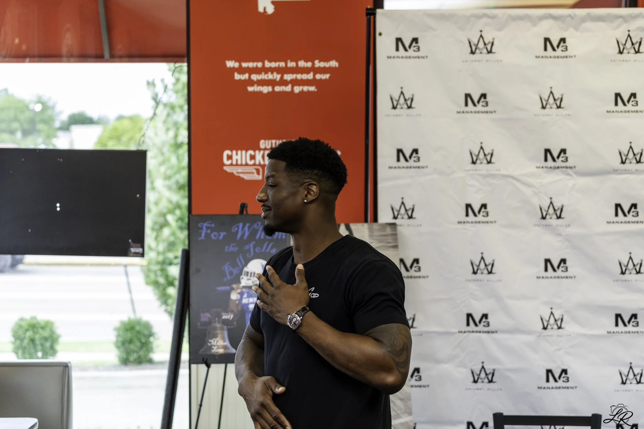 A man with short curly hair and earrings, wearing a black shirt, stands indoors with his eyes closed and hand on his chest. Behind him, there is a white patterned backdrop with a logo and the letter M repeated, and a red sign with white text saying, 
