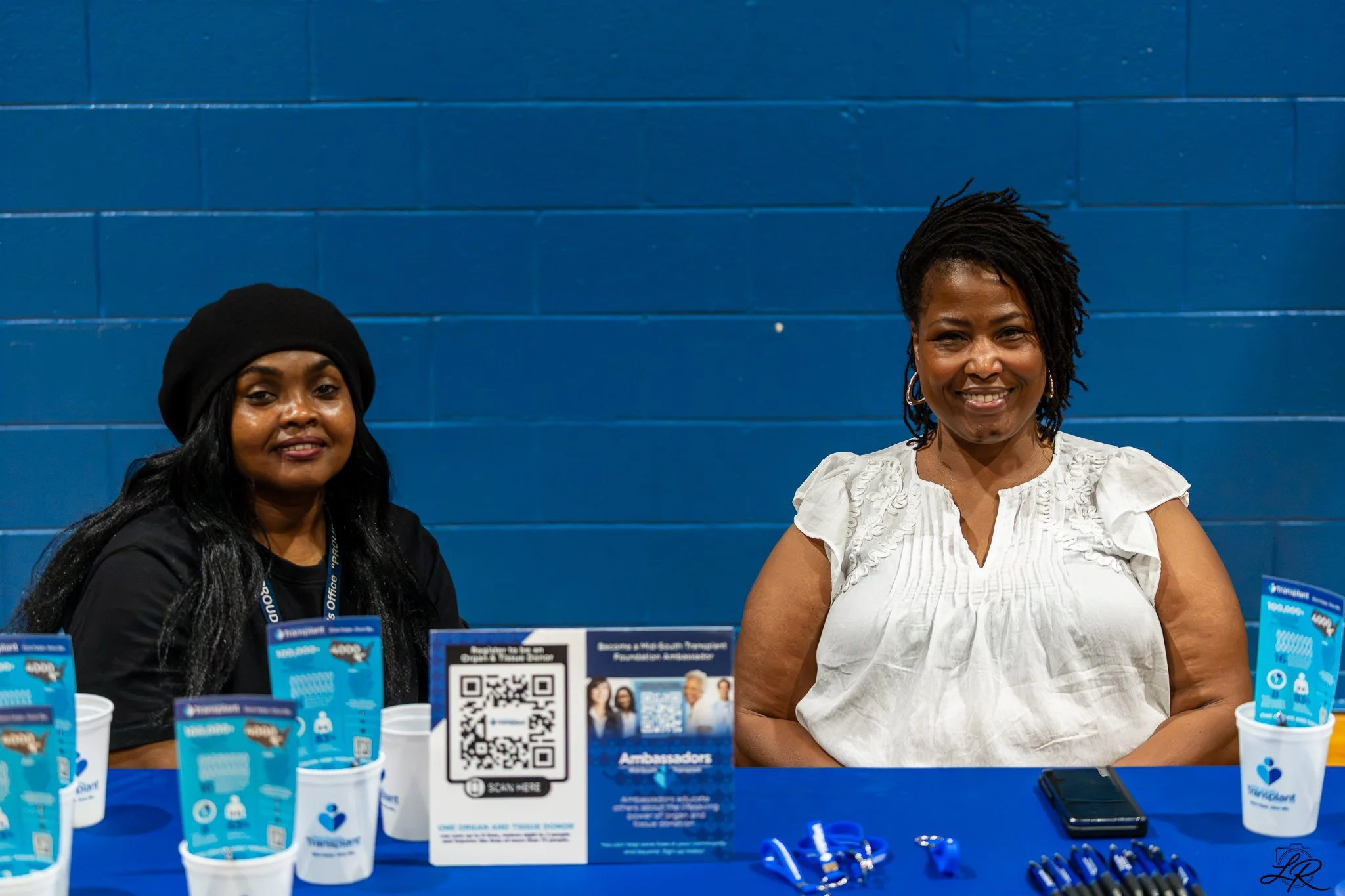 Two women sitting at a table with blue informational materials. The woman on the left wears a black hat and has long dark hair, the woman on the right has short curly hair and wears a white blouse. There is a blue wall behind them, and various promot