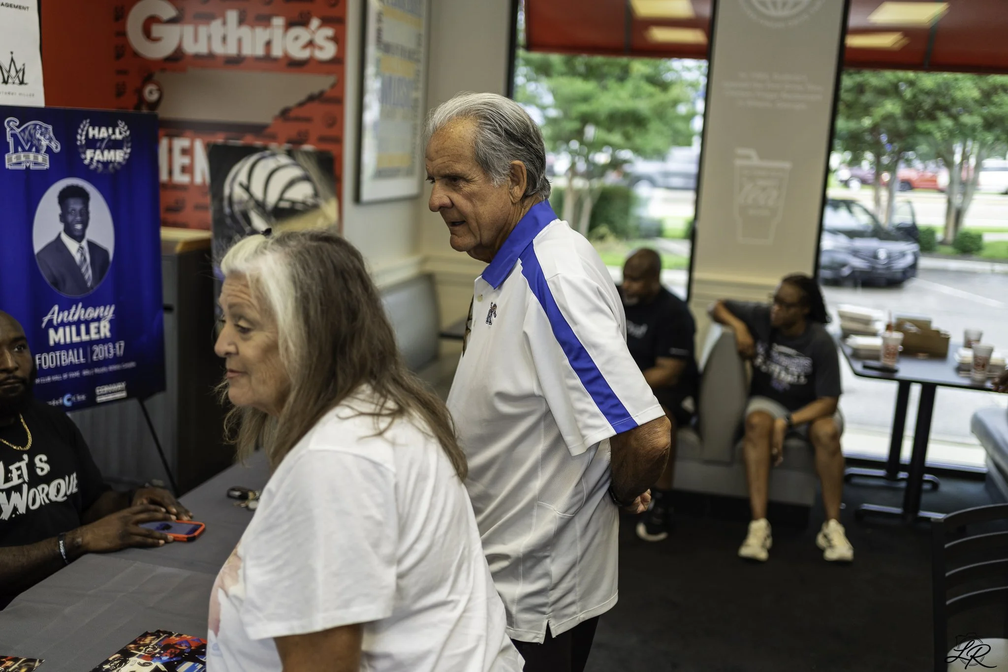 A man with gray hair wearing a white and blue shirt, talking to a woman with long gray hair at a meet and greet event at a restaurant. The background shows posters, people sitting and standing, and a window looking outside.