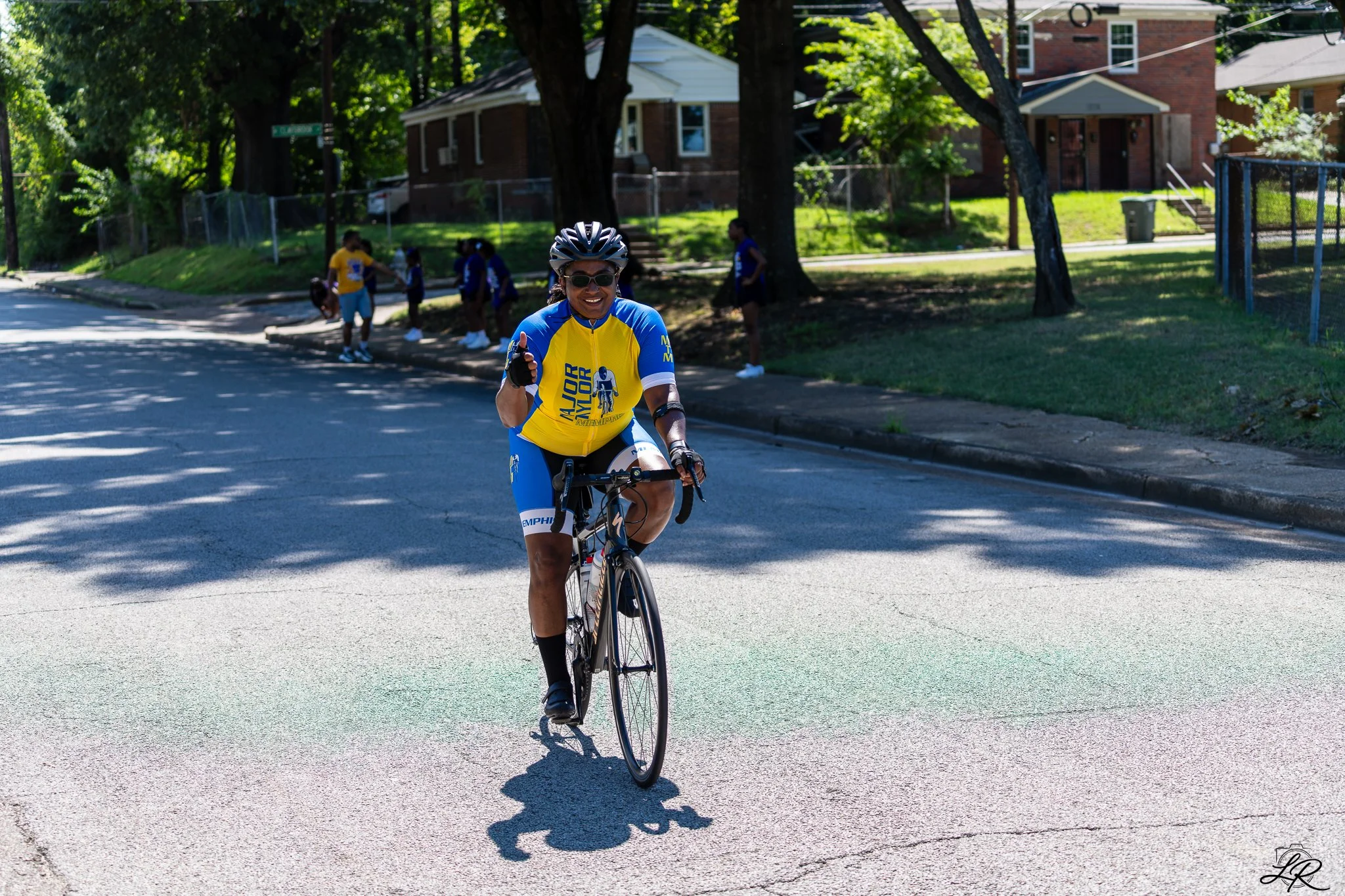 A woman wearing a blue and yellow athletic jersey, sunglasses, and a helmet riding a bicycle on a residential street during the daytime. She is smiling and giving a thumbs-up gesture. In the background, there are children standing under trees and hou