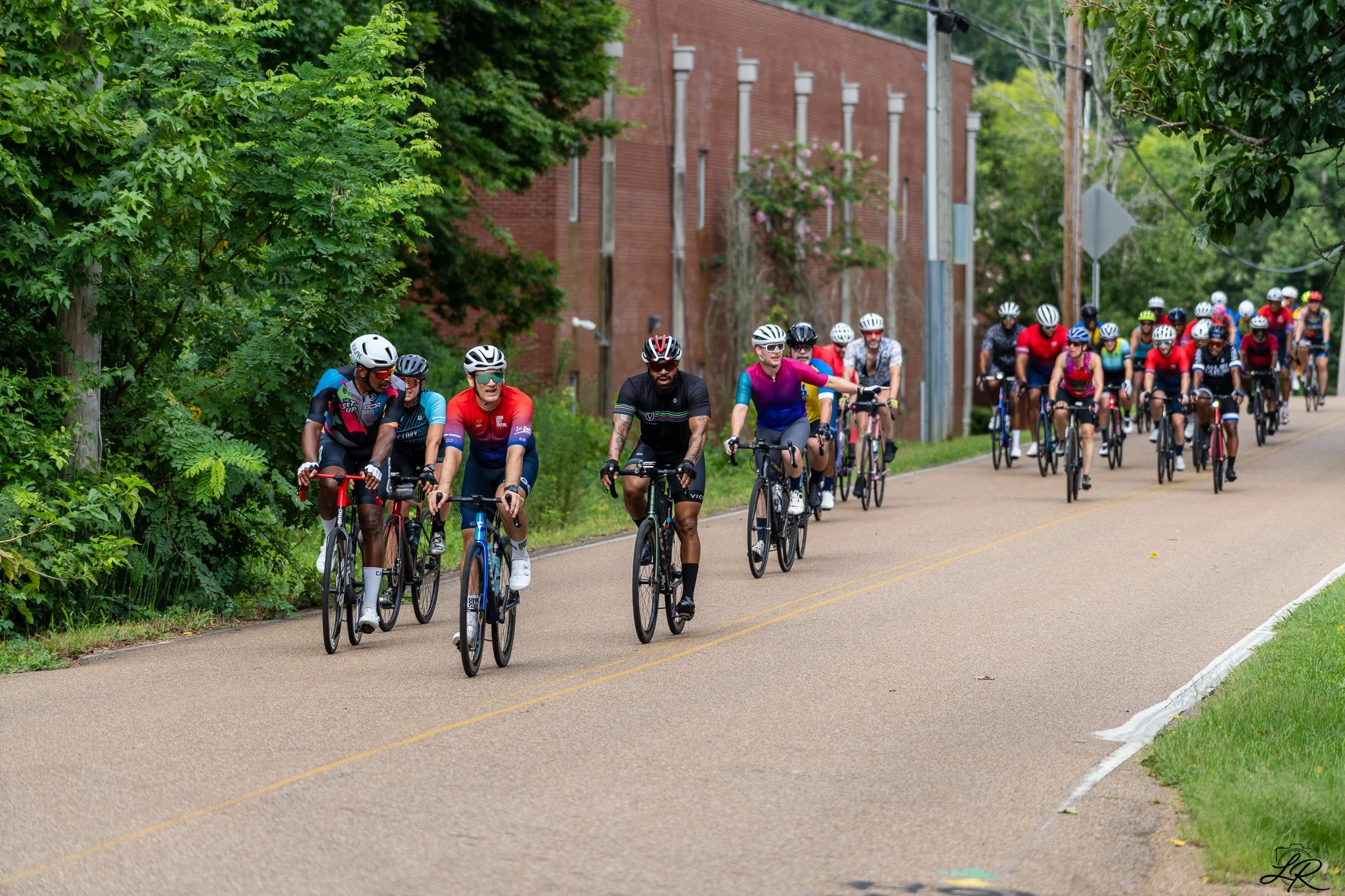 A large group of cyclists riding on a paved road surrounded by lush green trees and bushes, with a brick building and utility poles in the background.