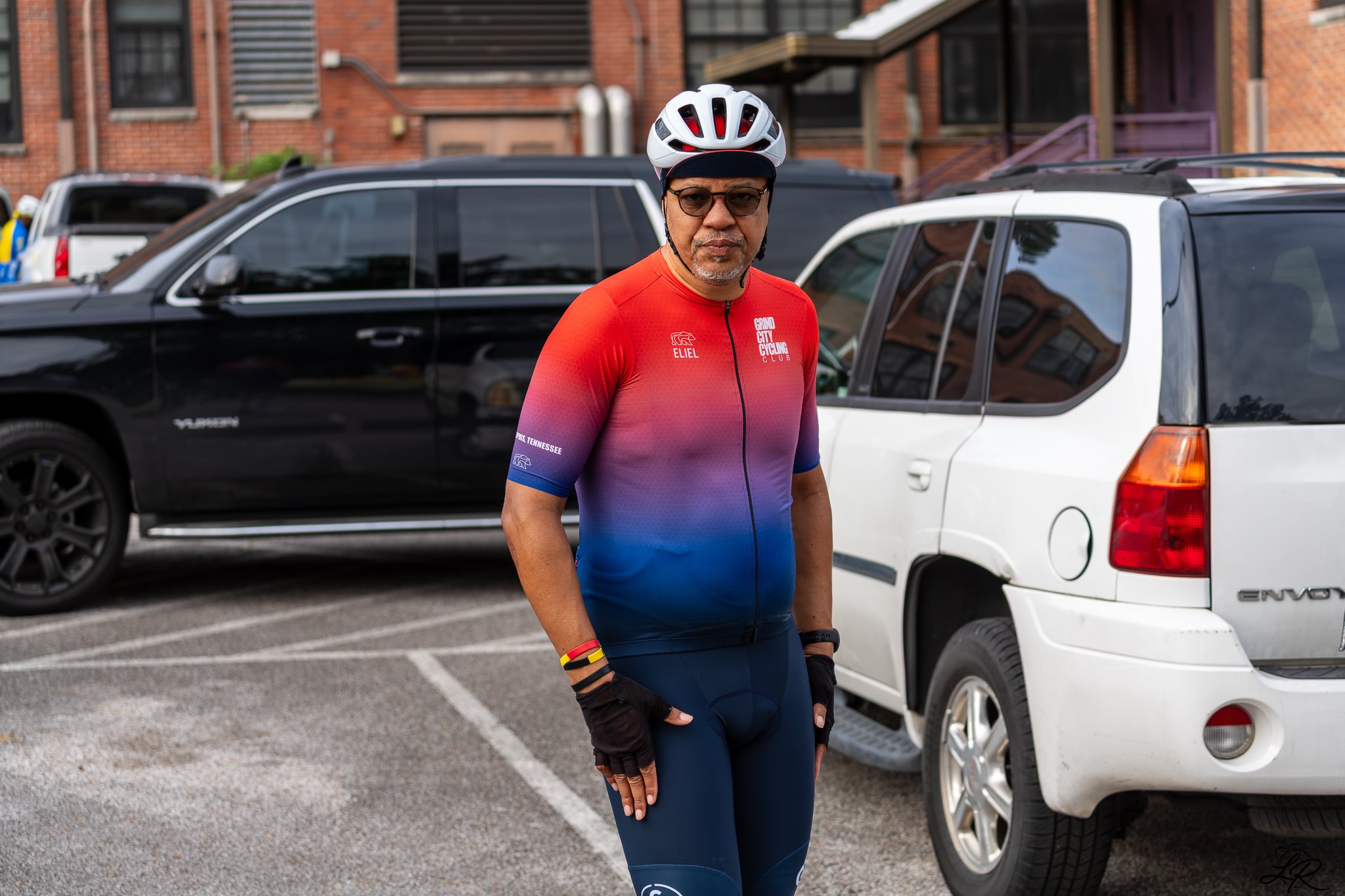 A man wearing a cycling helmet, sunglasses, and a colorful cycling jersey stands in a parking lot with cars behind him.