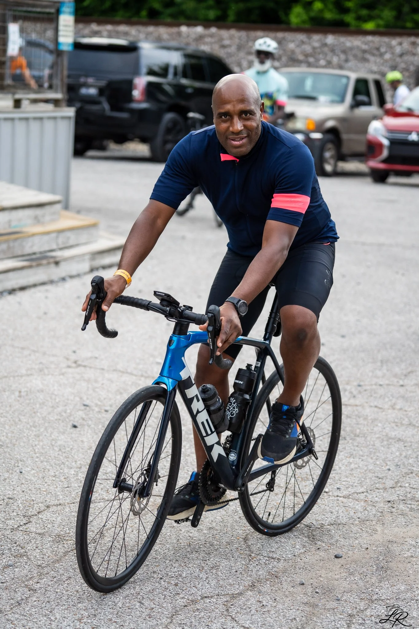 A man riding a bicycle on a gravel surface with parked cars and people in bicycle helmets in the background.