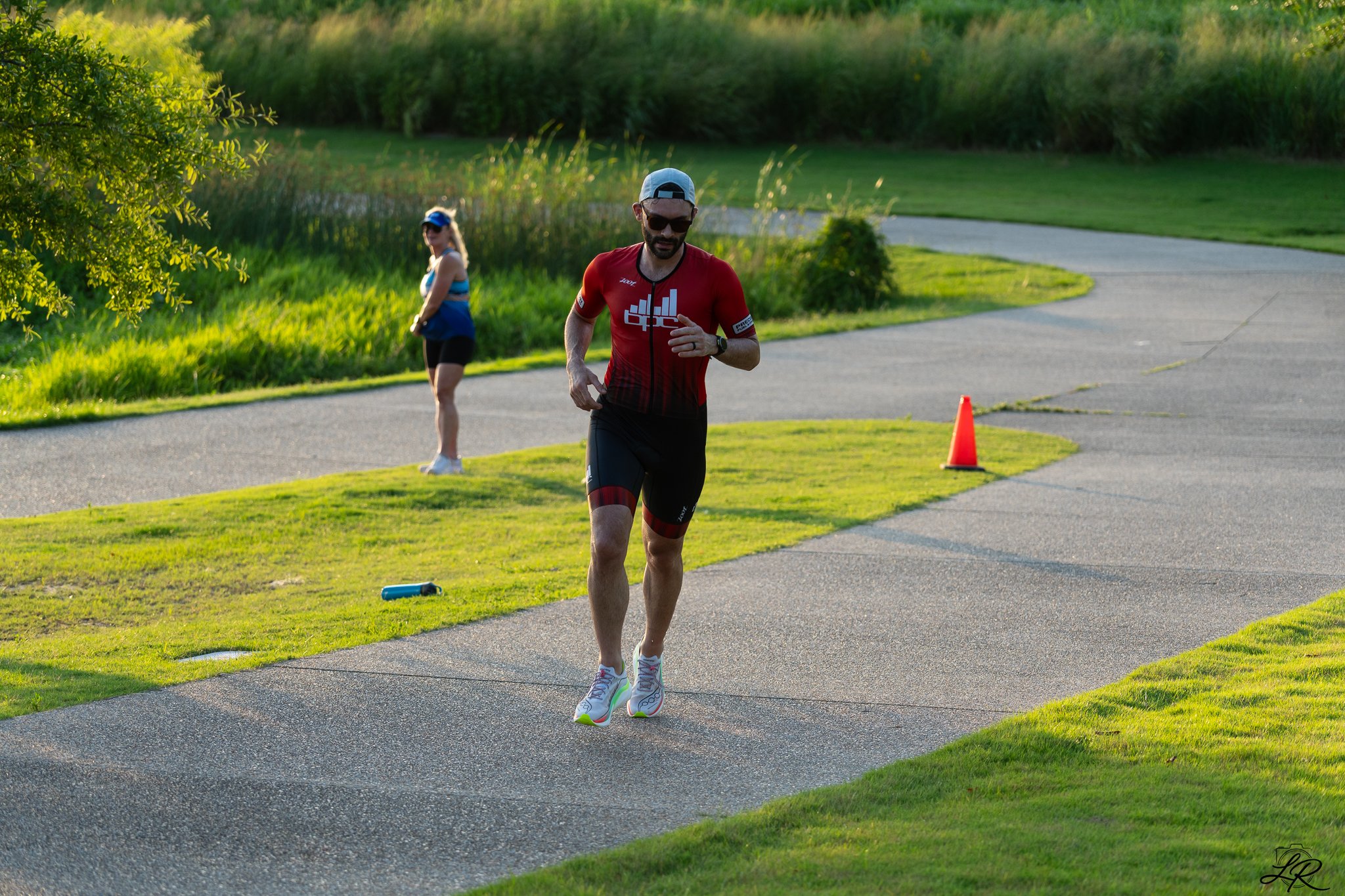 A man wearing sports attire, sunglasses, and a cap jogging outdoors on a paved path in a green park area. There is a woman in the background walking and a bright orange cone placed on the path.