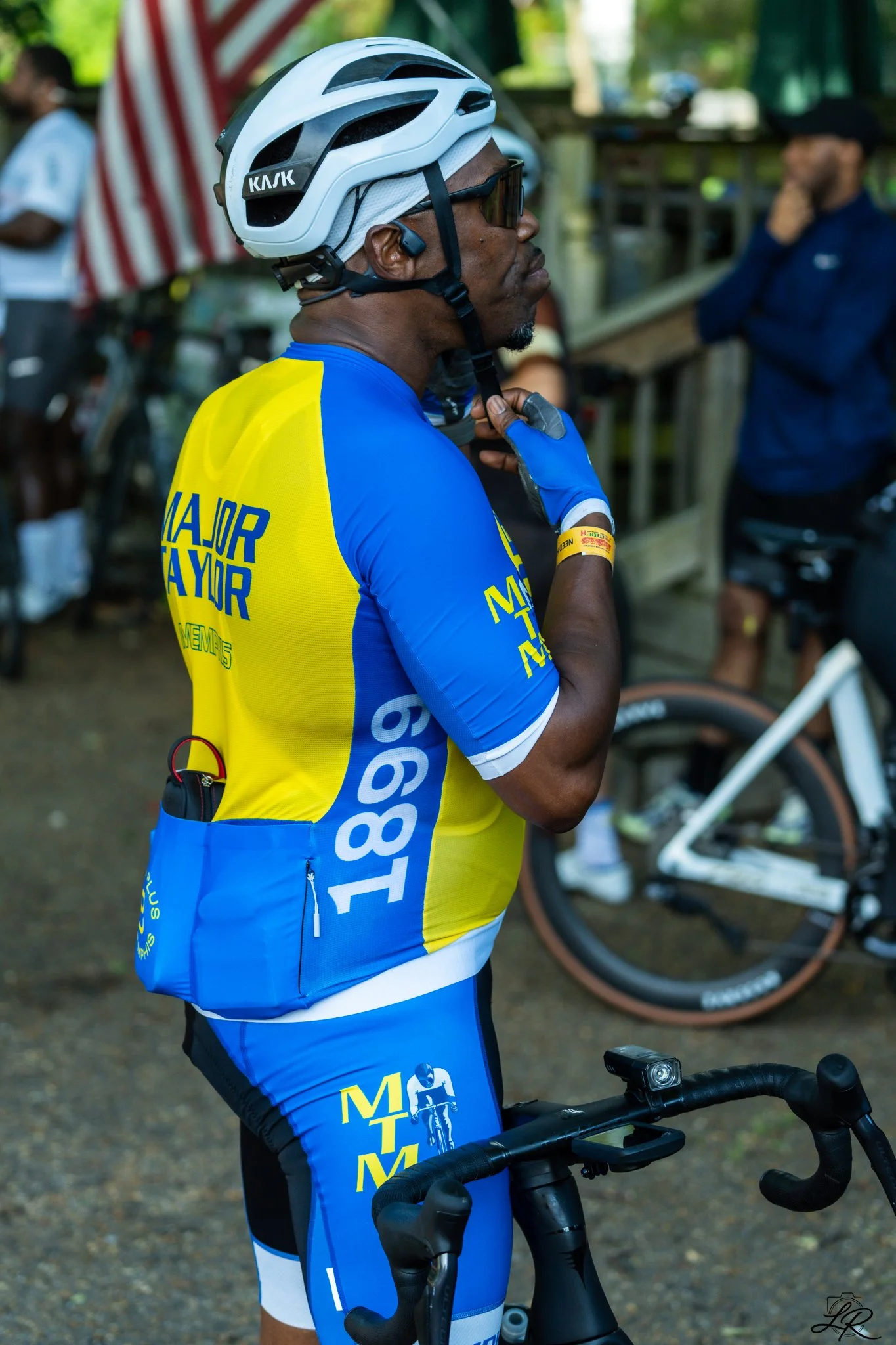 A man in cycling gear adjusting his helmet while holding the handlebars of a bicycle, with other people and a bicycle in the background.