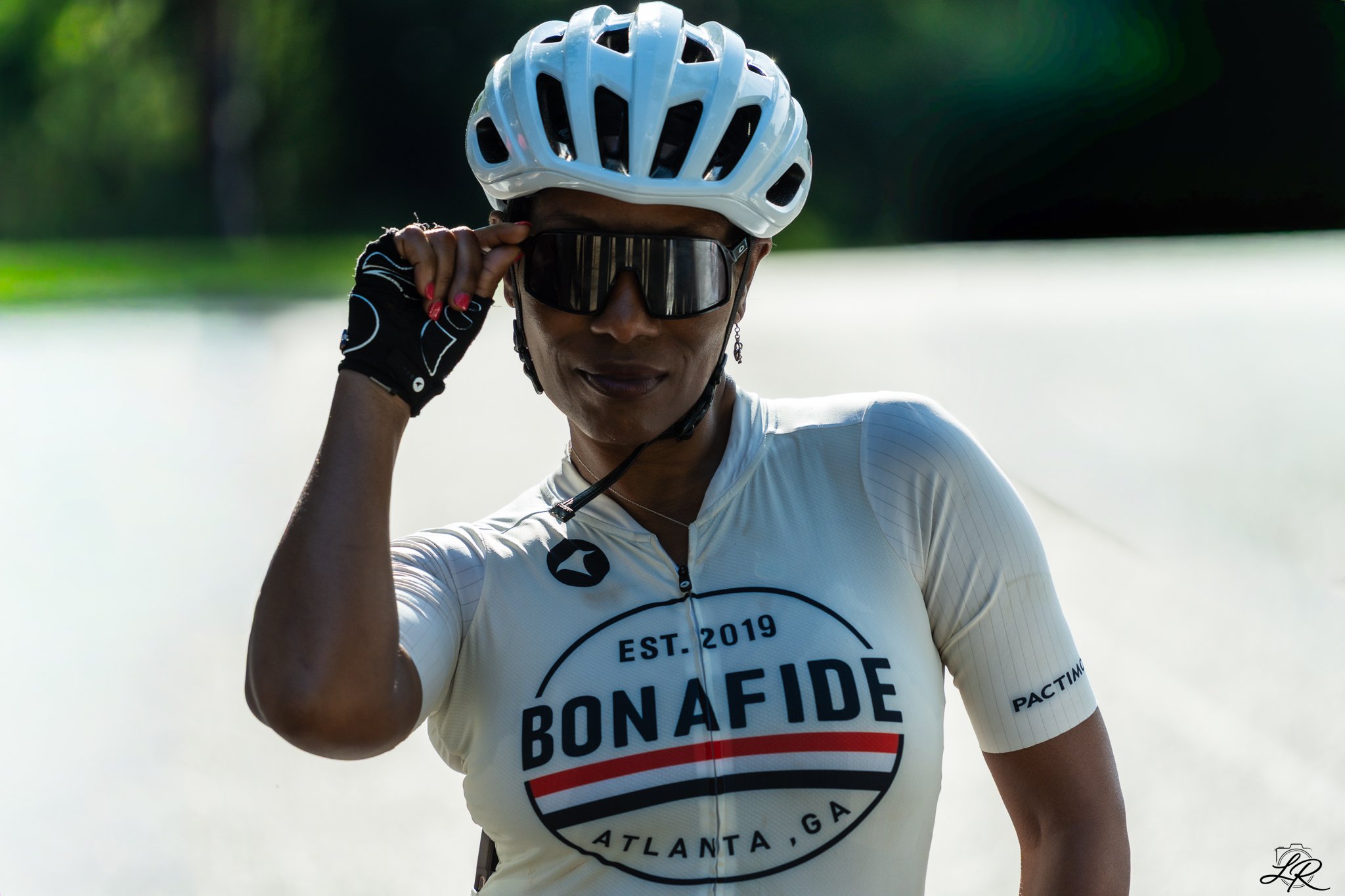 Close-up of a woman wearing a white cycling helmet and sunglasses, adjusting her glasses, dressed in a white cycling jersey with a logo, standing outdoors with a blurred background of water and greenery.
