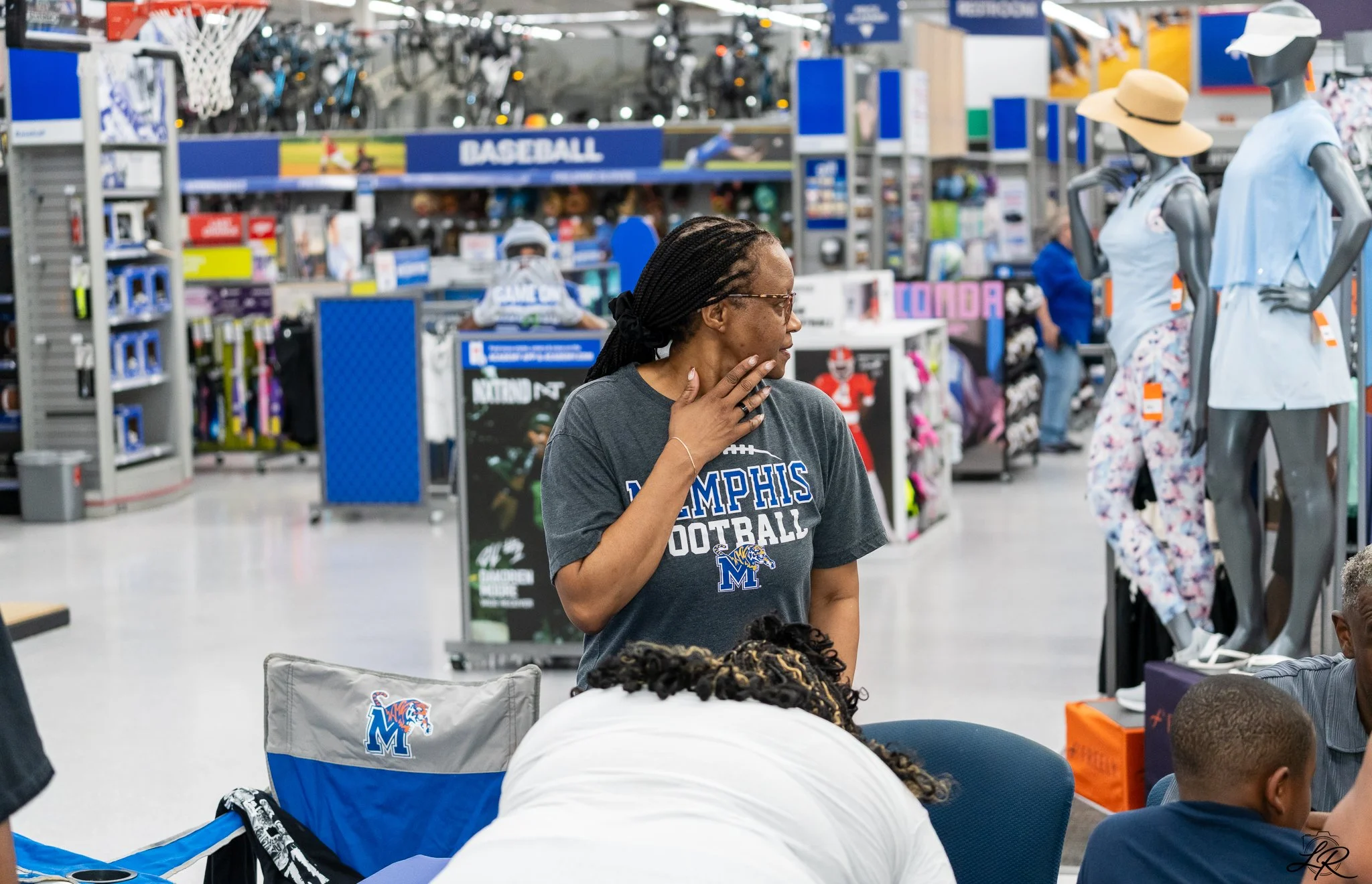 A woman with braided hair and glasses wearing a Memphis football t-shirt standing in a store aisle with mannequins dressed in summer outfits and hats, surrounded by sports equipment and apparel.