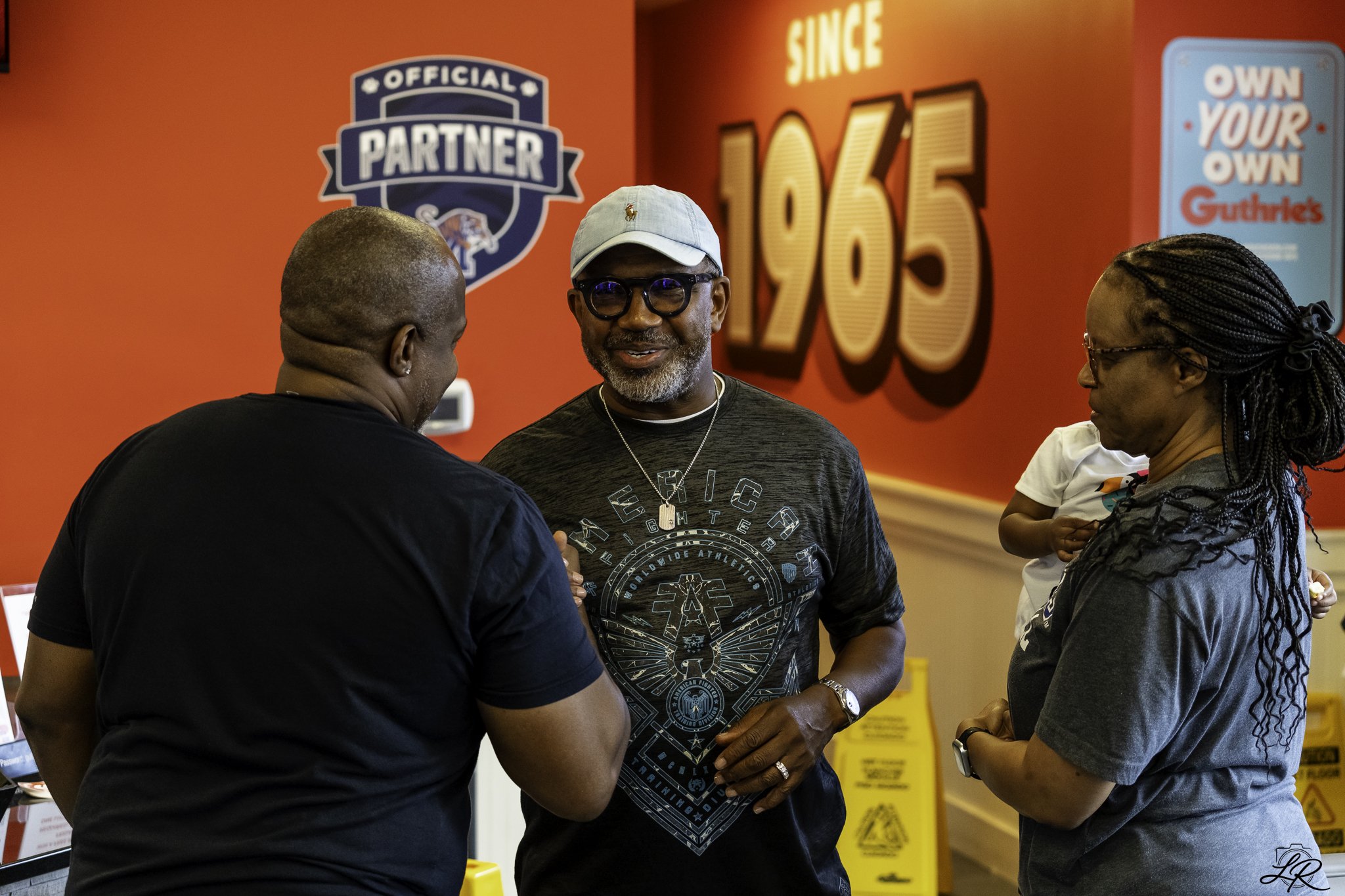 Four people standing together and talking in front of a red wall with branding and Olympic signage. The man in the middle is wearing glasses, a light gray cap, and a black sports shirt with a lion design and athletic branding. The other three are wom