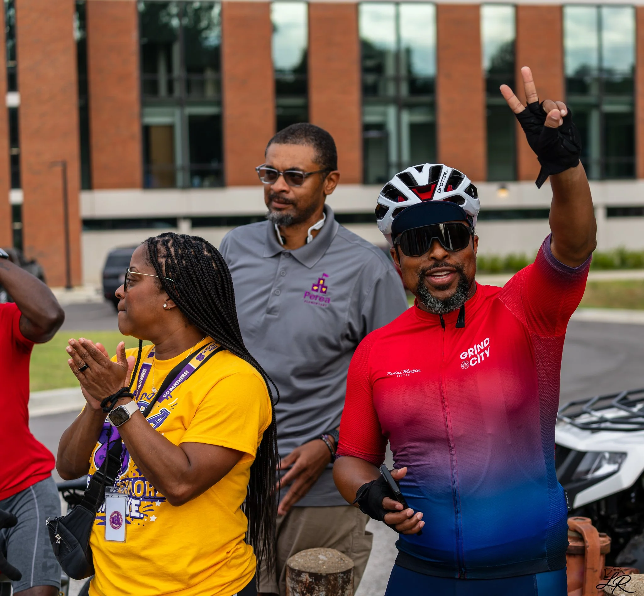 A group of diverse people gathered outdoors, some with hands raised or clasped, participating in an event or gathering. A woman in a yellow shirt with lanyards, a man in glasses, and others are visible, with a background of a modern brick building an